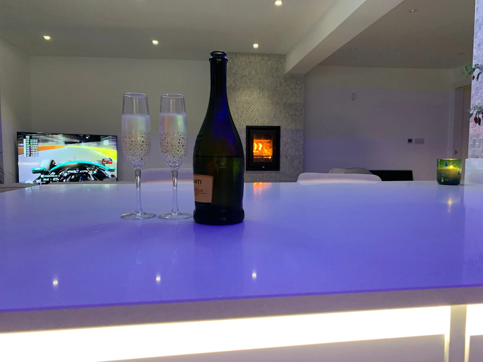 A bar counter with a bottle of champagne and two Champagne flutes, a television showing a racing game, a fireplace, and a green candle in a glass holder.