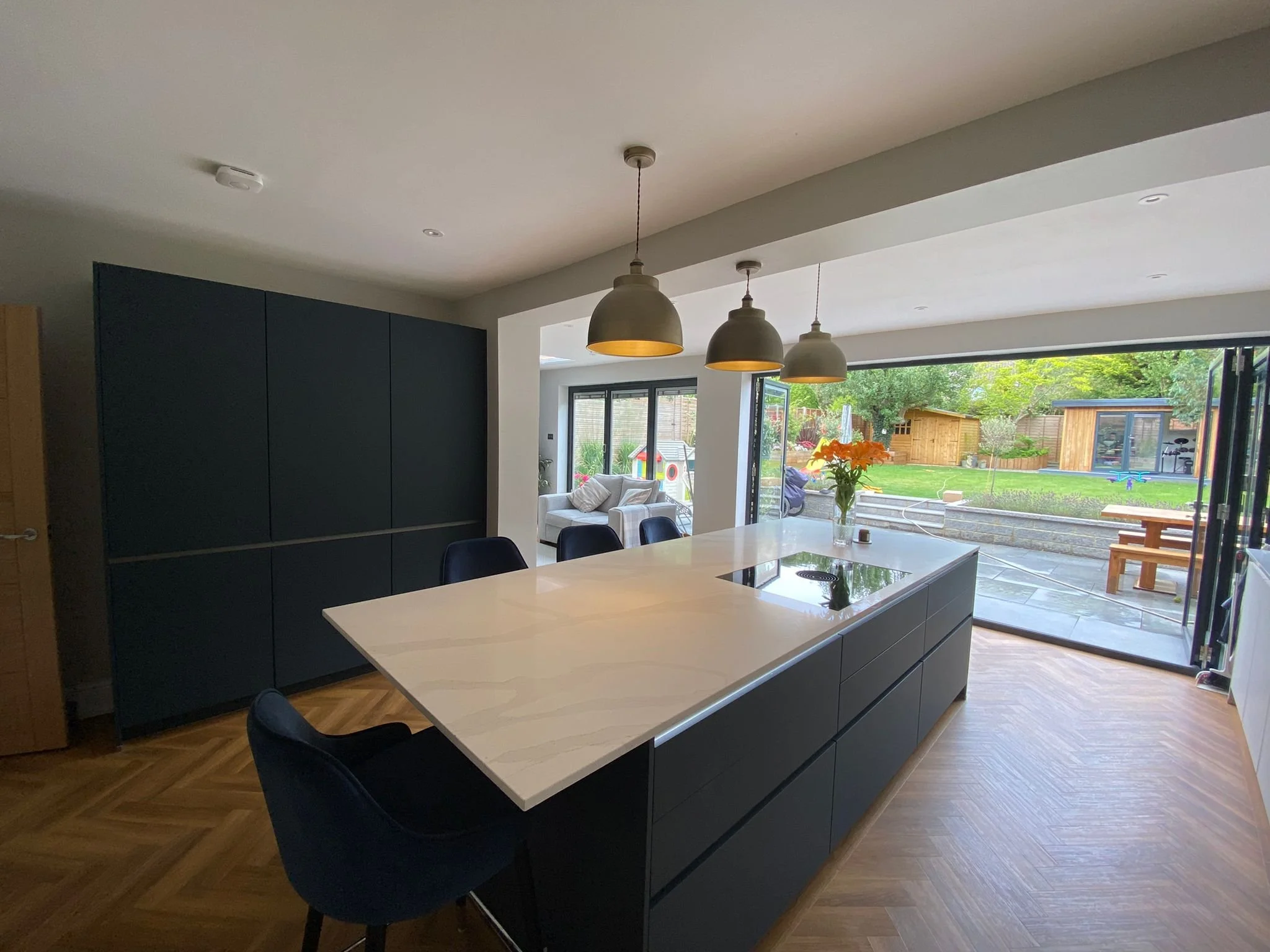Modern kitchen with an island, black cabinets, wooden flooring, and large glass sliding doors leading to a garden with outdoor seating.