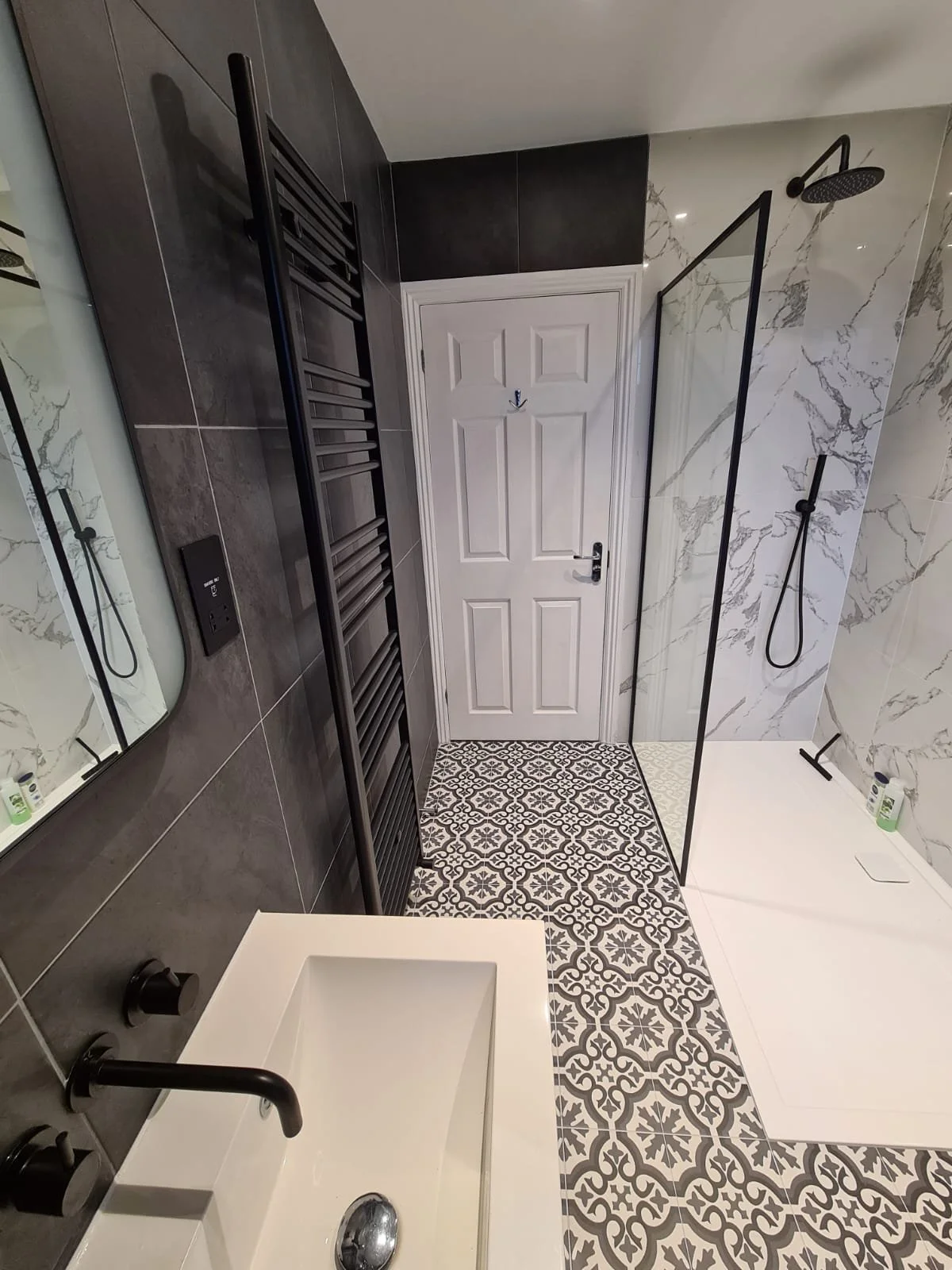 Modern bathroom with patterned floor tiles, a white door, a shower area with a black showerhead and a glass partition, a small white sink with black fixtures, a black towel radiator, and toiletries on the shower ledge.