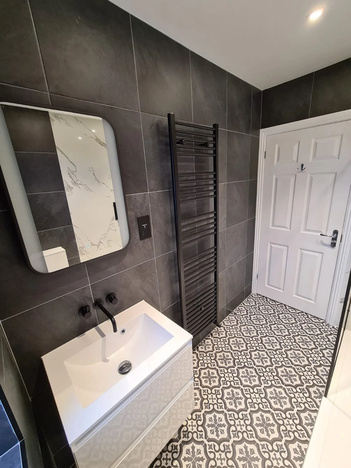 Modern bathroom with patterned tile floor, white sink with black fixtures, black towel heater, mirror, and white door with black hook.