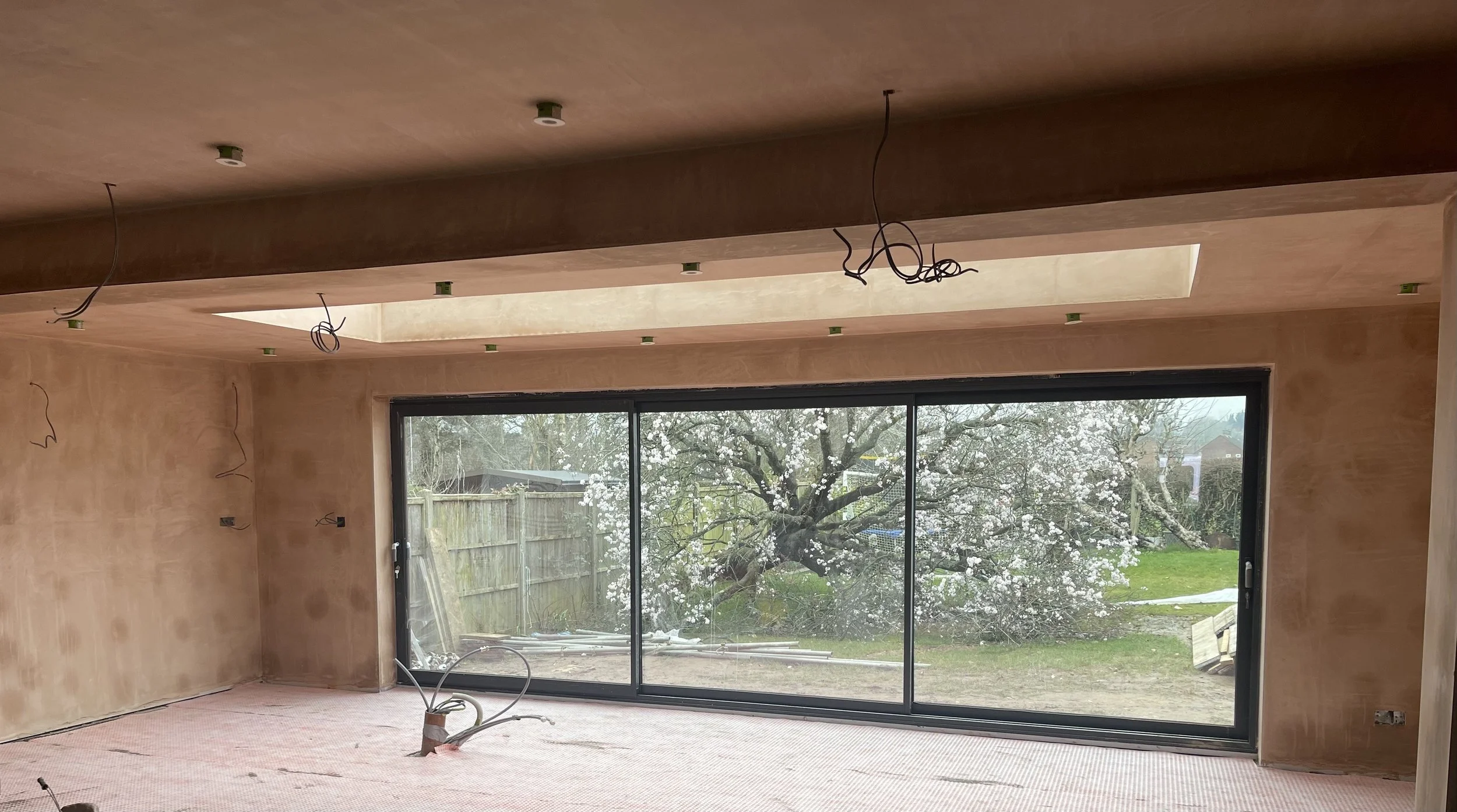 Interior of a room under construction with unfinished walls and ceiling. Electrical wiring hanging from the ceiling, large sliding glass door revealing a garden with blooming tree outside.