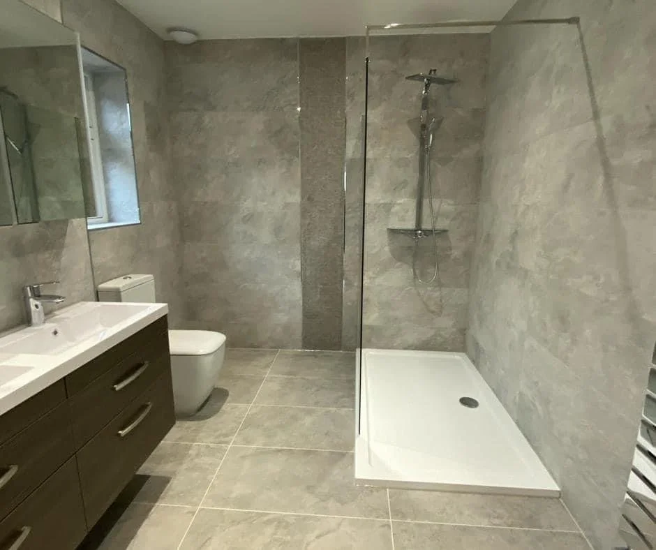 Modern bathroom with gray tiled walls and floors, a white toilet, a dark wooden vanity with a white sink, a wall-mounted mirror, a small window, and a glass-enclosed shower area with a showerhead and shelf.