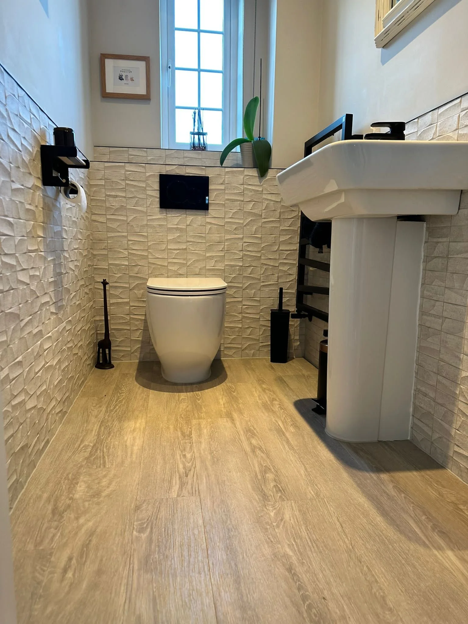 Small modern bathroom with beige tiled walls, a white toilet, a small white sink with black fixtures, a window with a potted plant, and a framed picture on the wall.