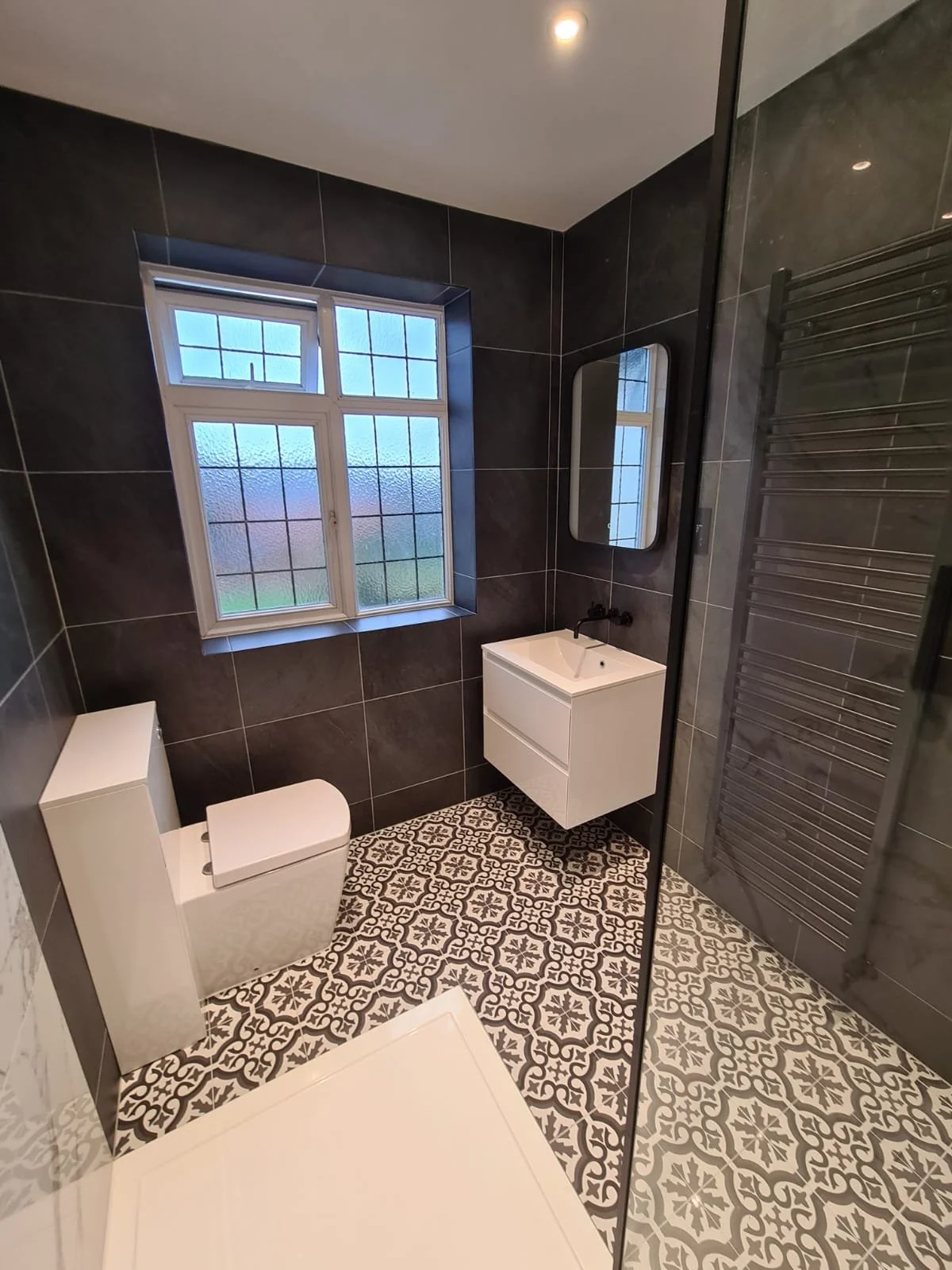 Modern bathroom with black tiled walls, patterned floor tiles, a white toilet, a small white vanity sink with a black faucet, a black-framed mirror, a window with frosted glass, and a heated towel rail.