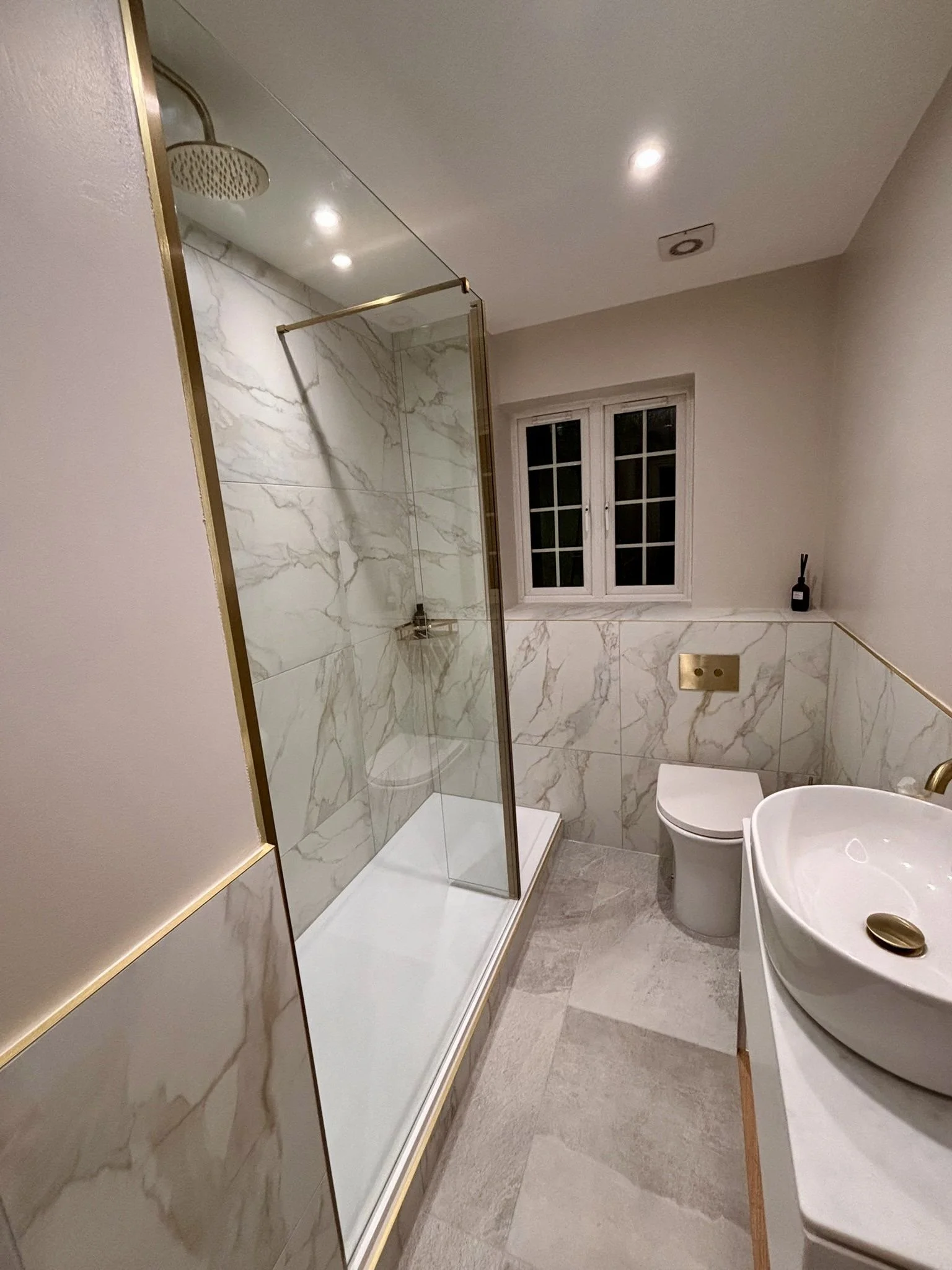 Modern bathroom with marble wall tiles, glass shower enclosure, white toilet, white vessel sink with gold fixtures, window, and neutral-colored flooring.