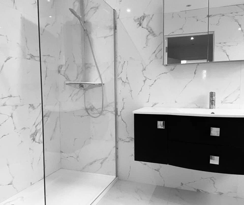 A modern bathroom with marble walls, a glass shower enclosure with a showerhead and shelf, a black floating vanity with a white countertop, a faucet, and a rectangular mirror.