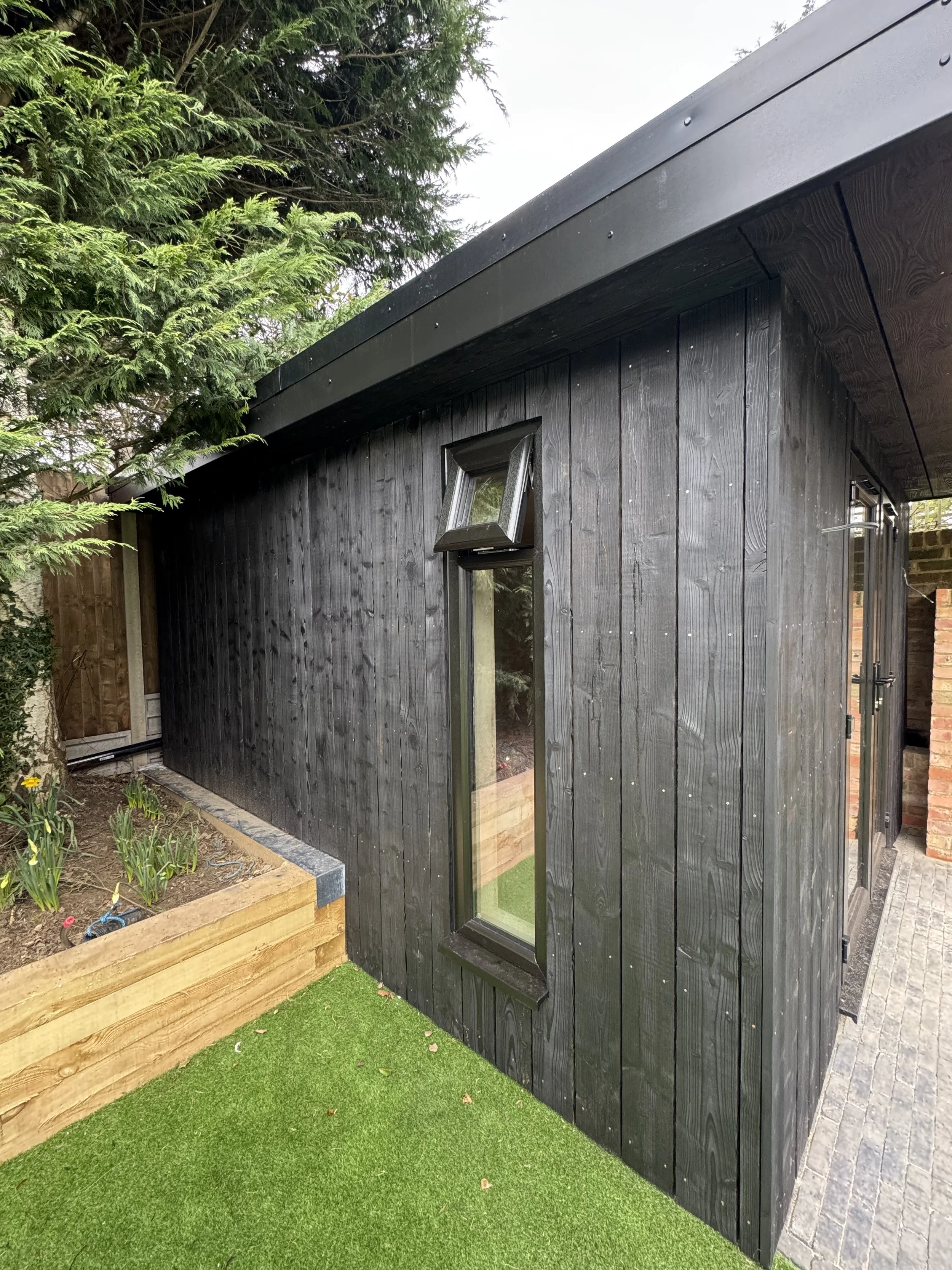 A small black wooden shed with a sloped roof, narrow window, and a small window vent. It is adjacent to a flower bed and a brick wall.