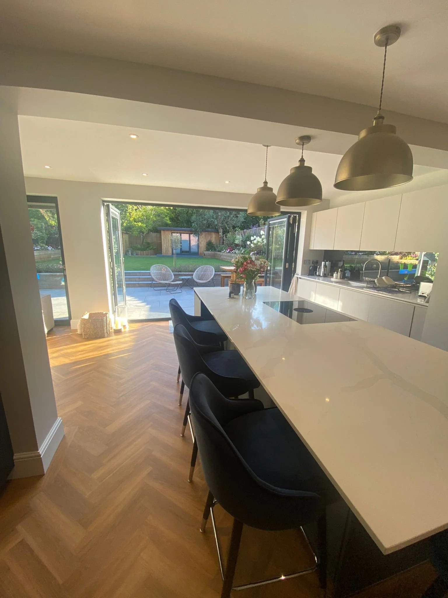 Modern kitchen with white cabinetry, a large island with black chairs, and a view into a backyard with a patio, garden, and outdoor sitting area.