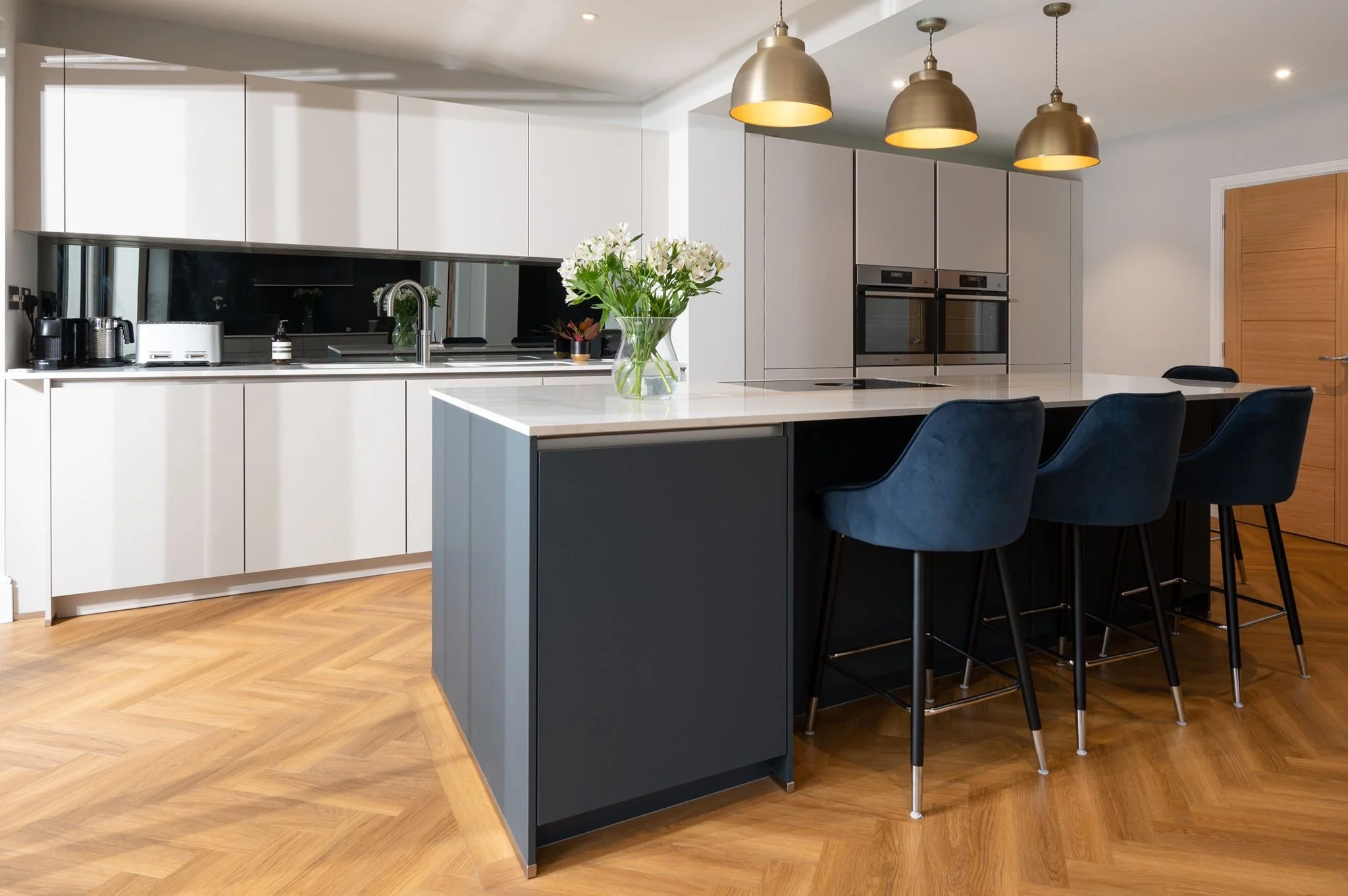 Modern kitchen with white and dark gray cabinetry, a marble island with dark blue barstools, hardwood flooring, pendant lights, a vase of white flowers on the island, and built-in stainless steel appliances.