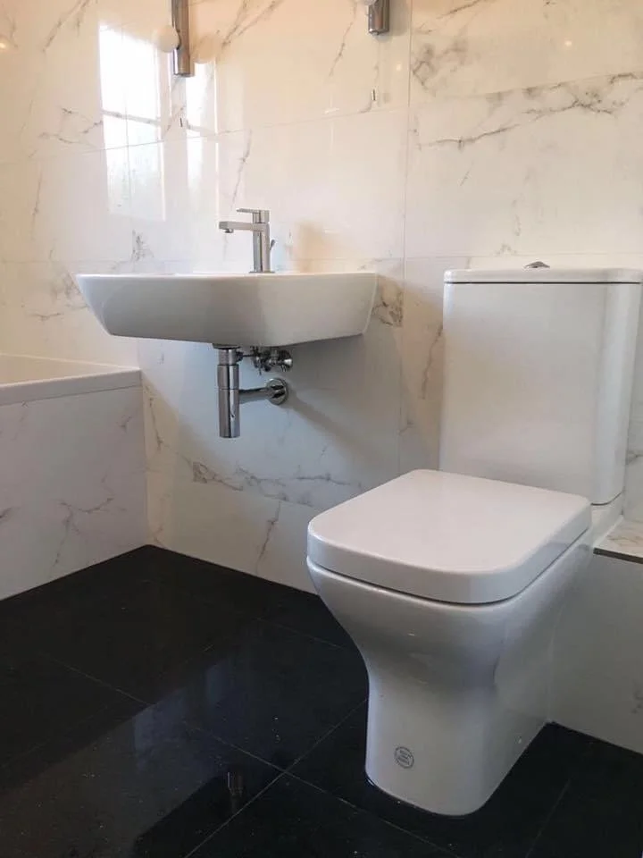 Bathroom with a wall-mounted sink and a toilet, marble walls, black tiled floor, and a window reflected in the mirror.