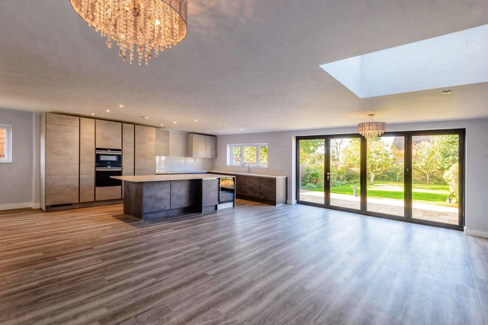 Modern, empty kitchen and dining area with large sliding glass doors opening to a green backyard, featuring wood flooring, light-colored cabinets, and a chandelier.