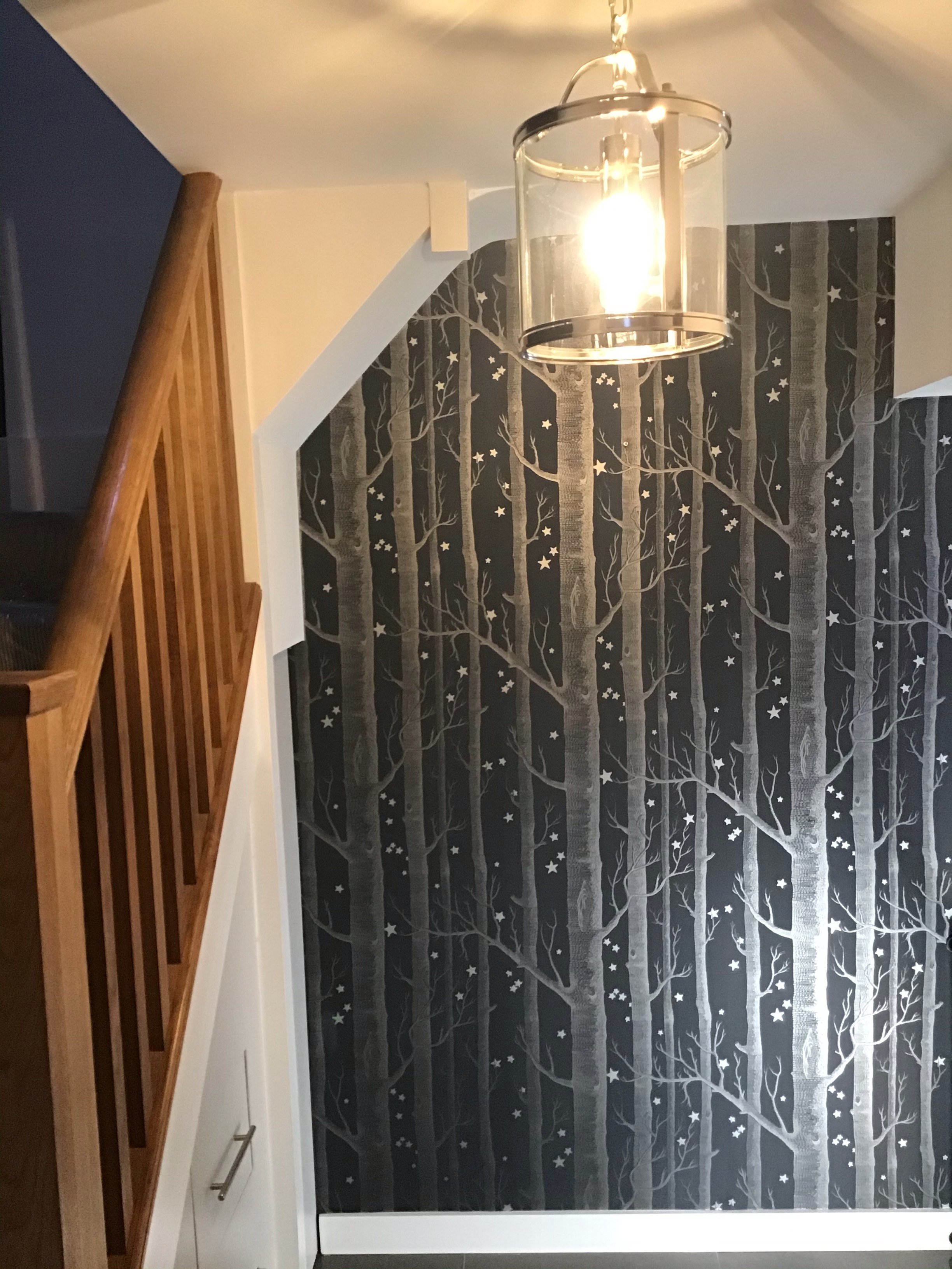 Interior view of a hallway corner with a staircase and a dark wall decorated with a tree-themed wallpaper with stars. A modern hanging ceiling light fixture is also visible.