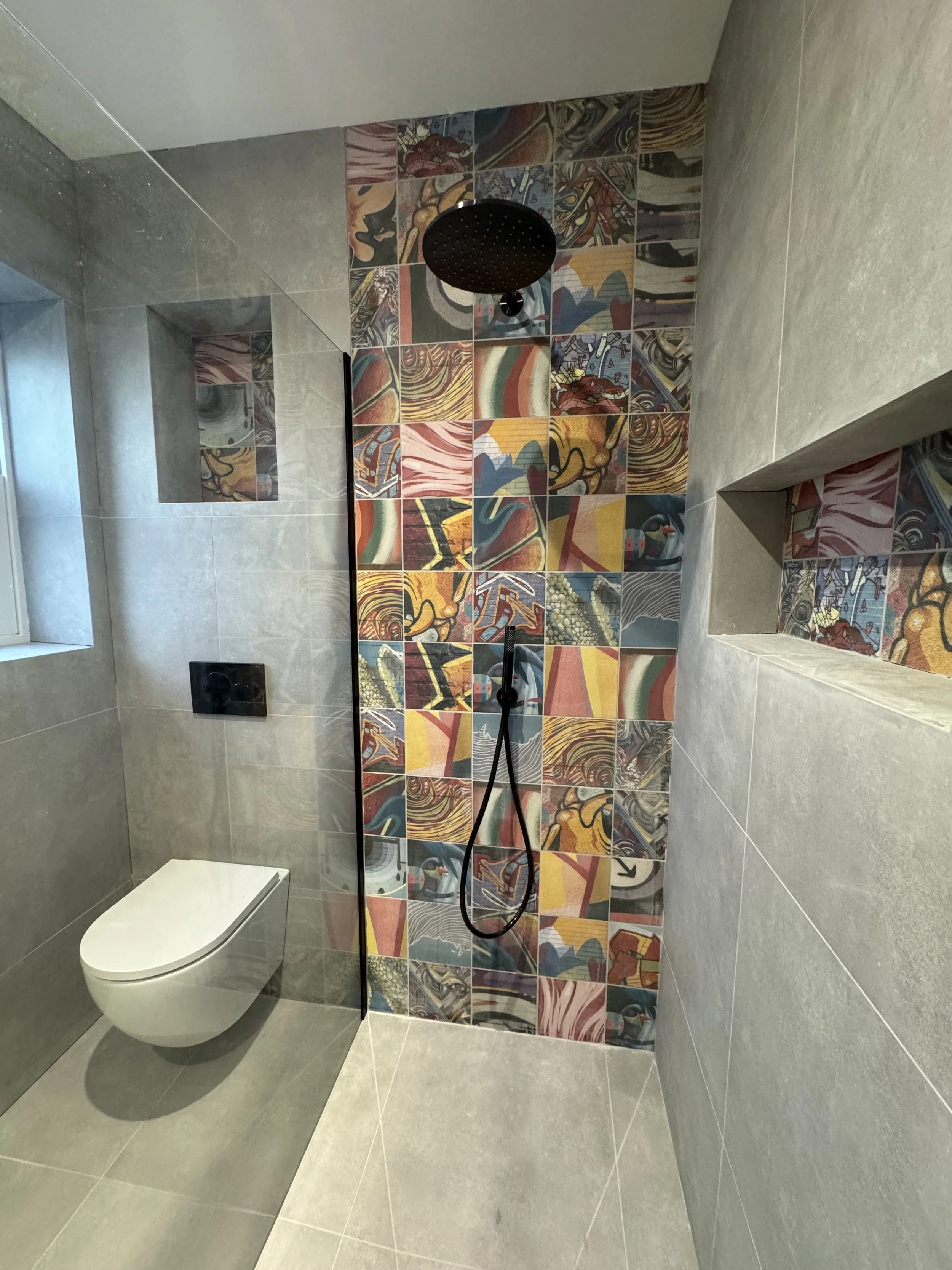 Modern bathroom with gray tiles and a colorful abstract tiled shower wall featuring a black rainfall showerhead and a handheld shower.