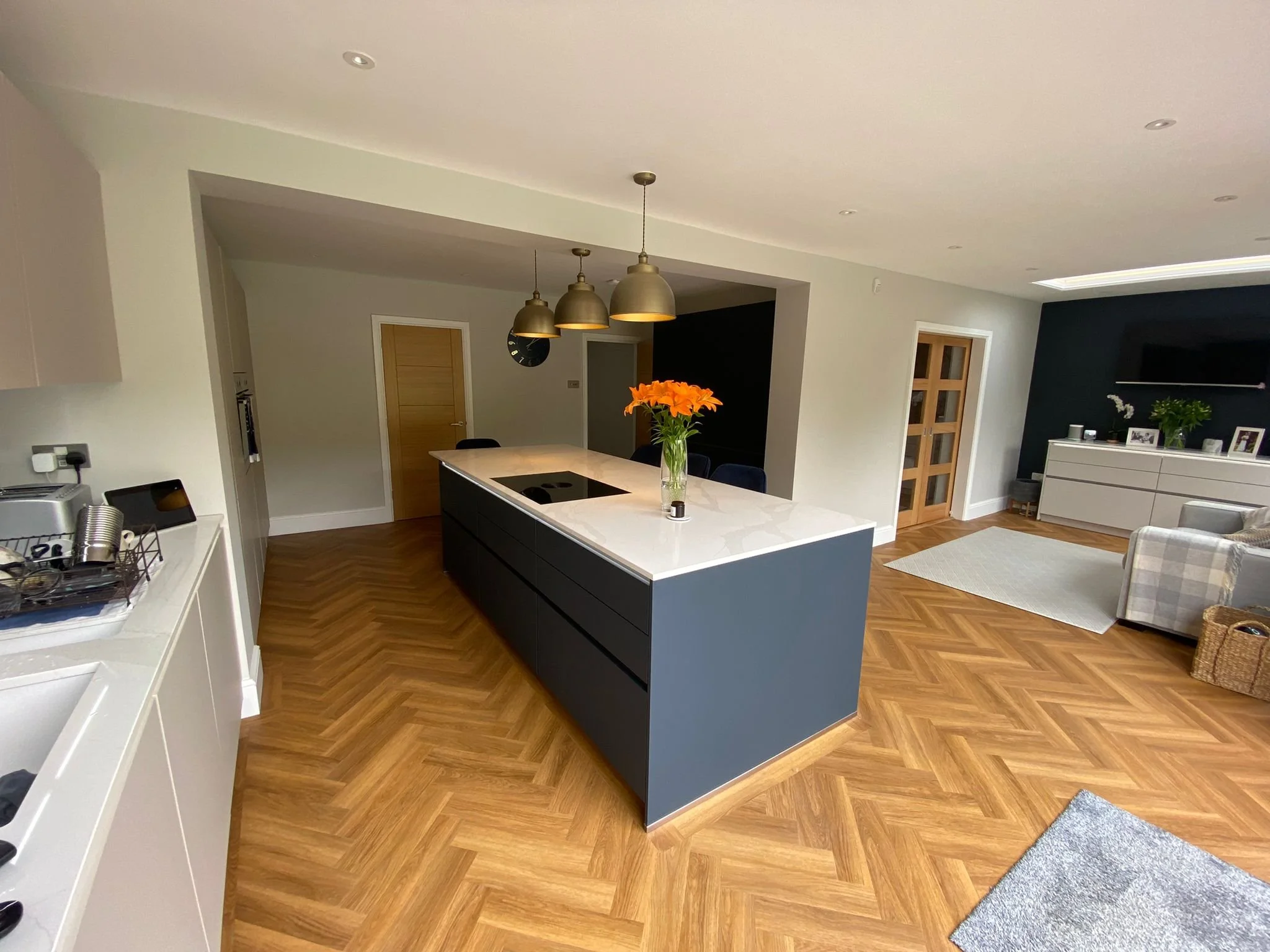 Modern open-plan kitchen and living room with wooden flooring, kitchen island with white countertop, gray and beige furnishings, and floral decorations.