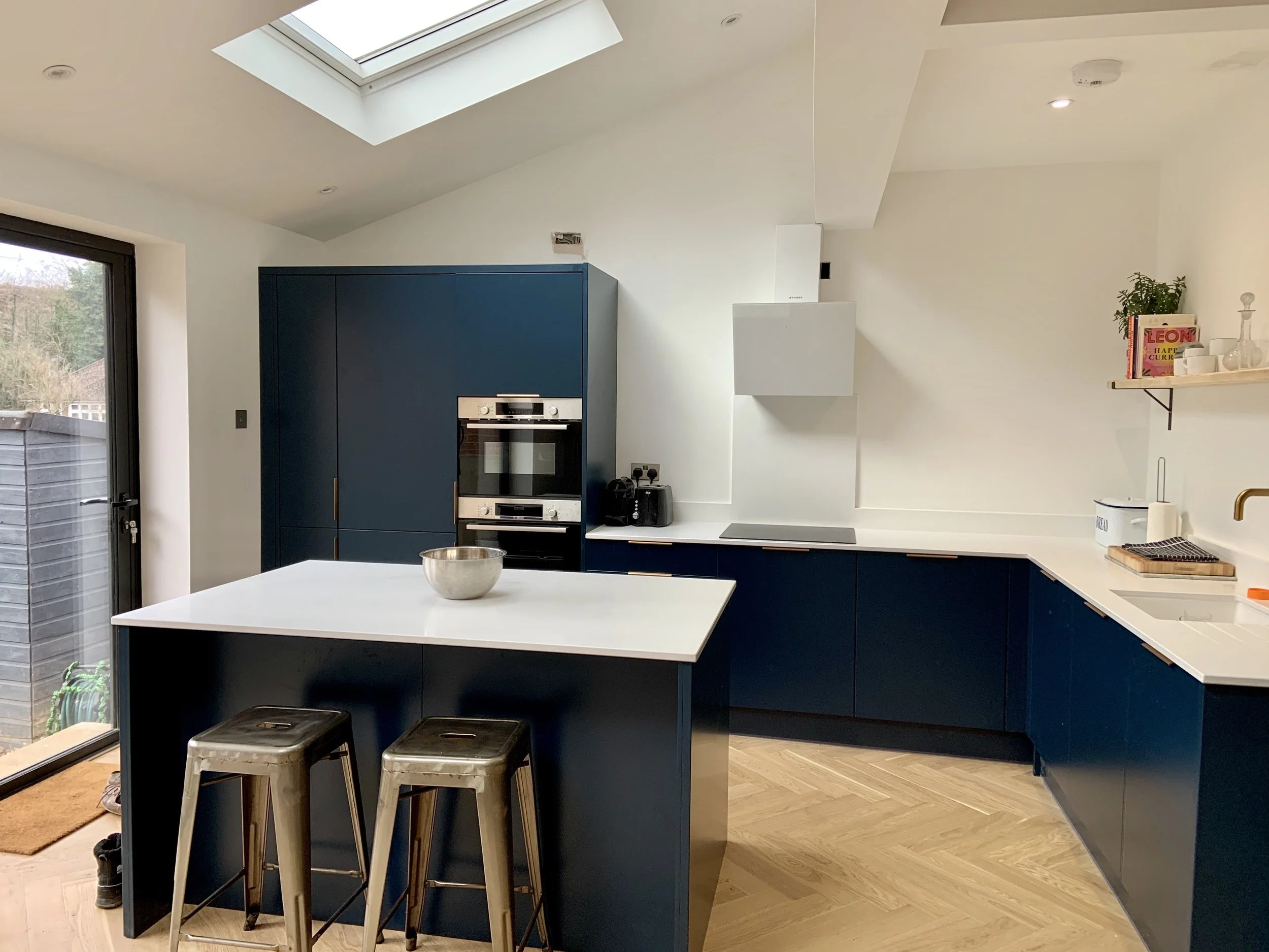 Modern kitchen with navy blue cabinets and white countertops, an island with two metal stools, built-in oven, stovetop, skylight, sliding glass door, wooden flooring, and a black shelf with plants, books, and kitchenware.