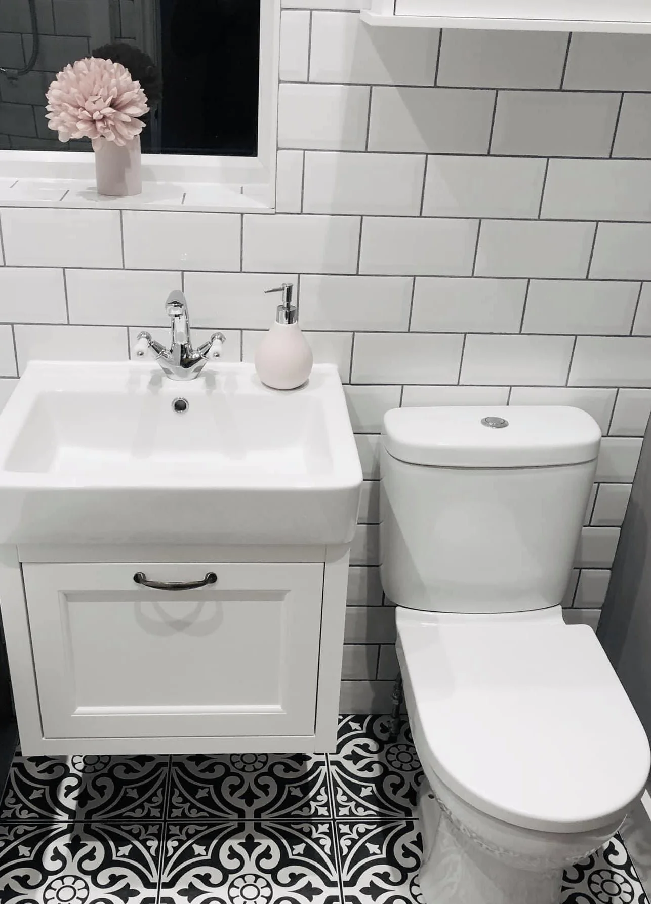 Bathroom with a white sink cabinet, a faucet, a soap dispenser, a toilet, and a window with a pink flower in a vase on the windowsill.