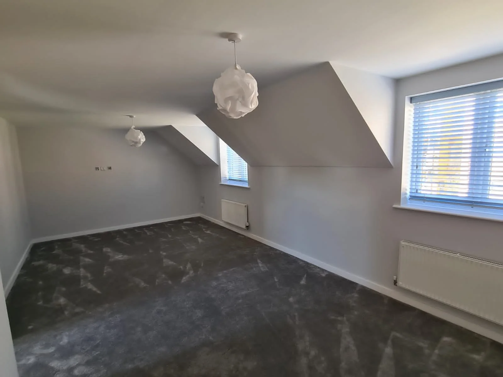 Empty room with gray carpet, white walls, two windows with blinds, and two hanging light fixtures.