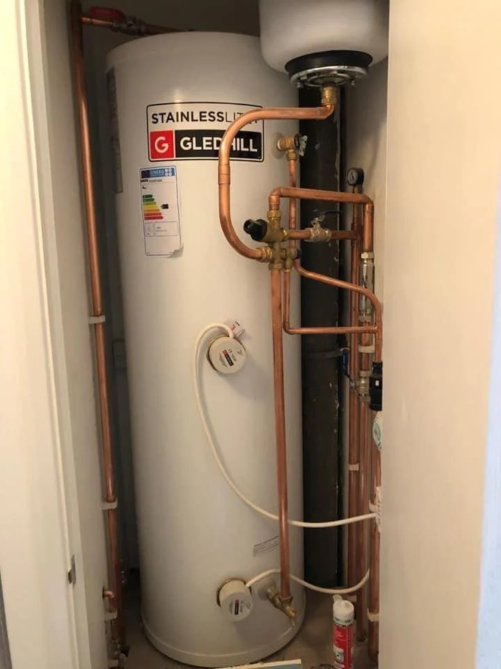 A white Gledhill stainless steel water heater with copper pipes connected to it, installed in a small closet space.