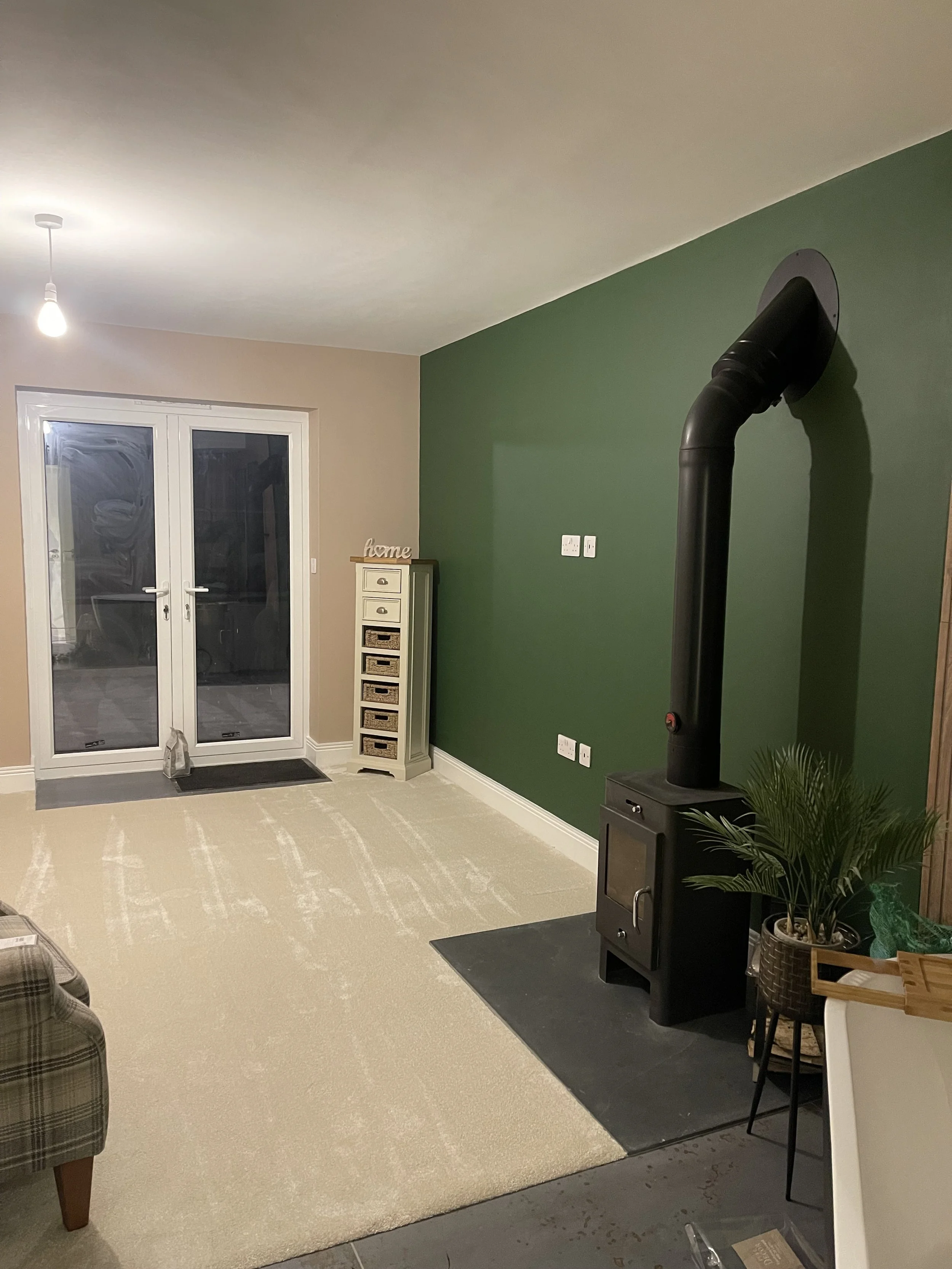 Living room with beige carpet, green accent wall, white sliding glass doors, a small white chest of drawers, a wood sign that says 'home', a black wood stove with a chimney pipe, and a potted plant.