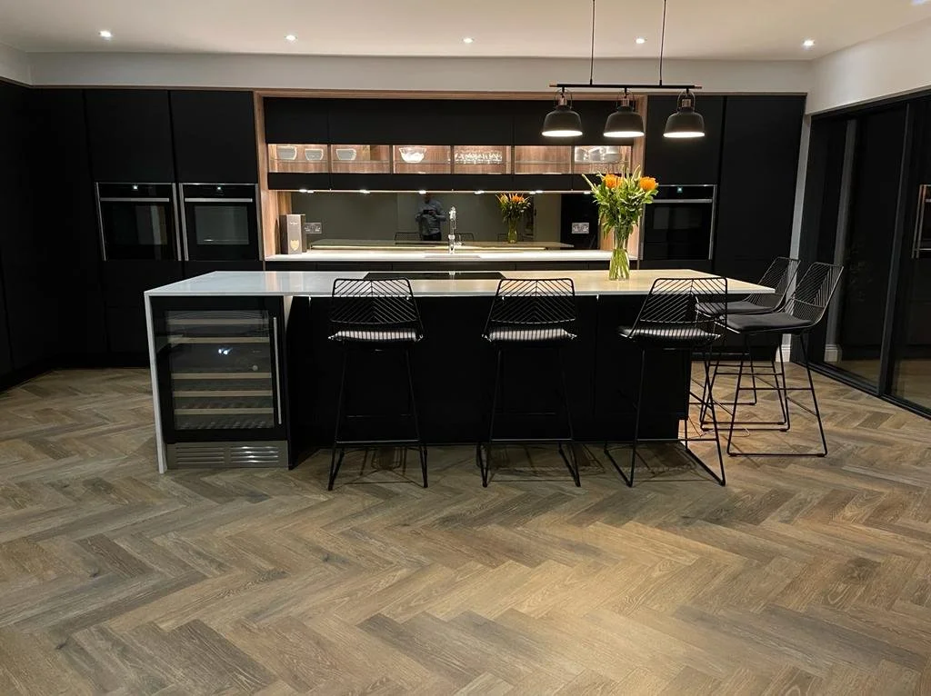 Modern black kitchen with a white countertop island, black barstools, and a wine cooler, featuring pendant lighting and floral arrangements.