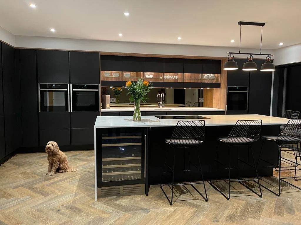 Modern black kitchen with a white island counter, bar stools, a built-in wine cooler, and a vase of flowers, with a small dog sitting on the wooden floor.