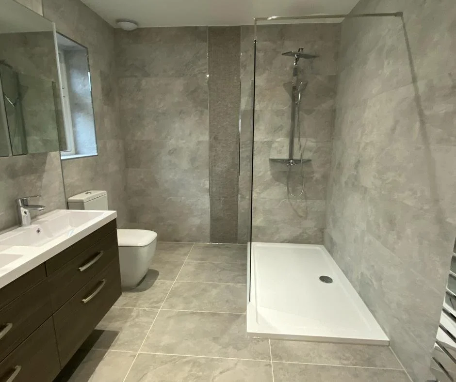 Modern bathroom with gray tiled walls and floor, a white shower tray with a glass partition, a dark wood vanity with a white sink, a toilet, and a small mirror cabinet.