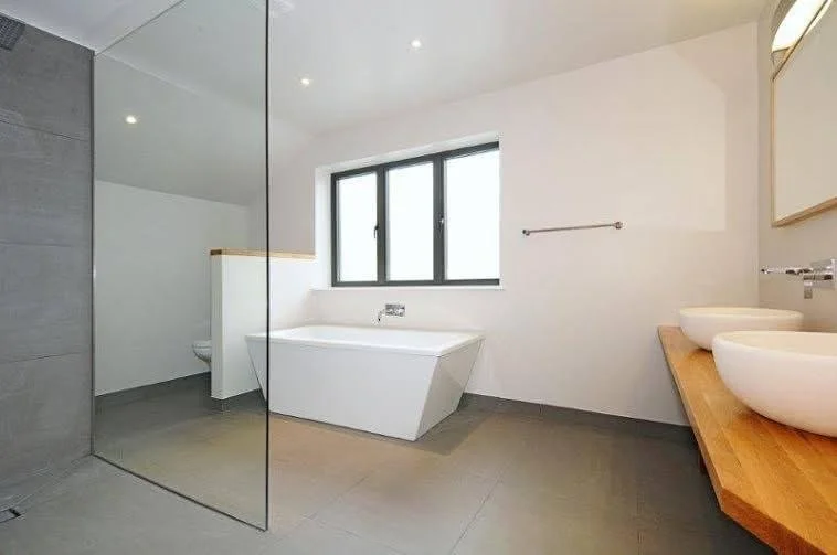Modern bathroom with a white bathtub beneath a large window, a wooden countertop with two vessel sinks, and a glass shower enclosure, all with minimalist design and neutral colors.
