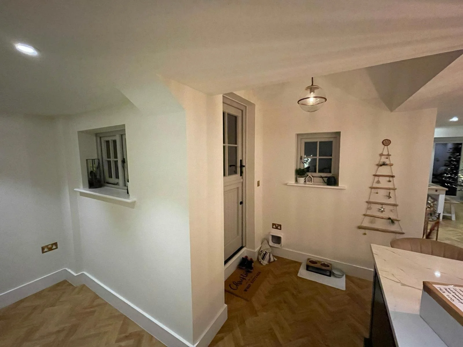 Interior view of a cozy home entryway with a small window, a wall-mounted wooden Christmas tree, a potted plant, and a door leading outside. The flooring is light wood, and there are warm ambient lights.