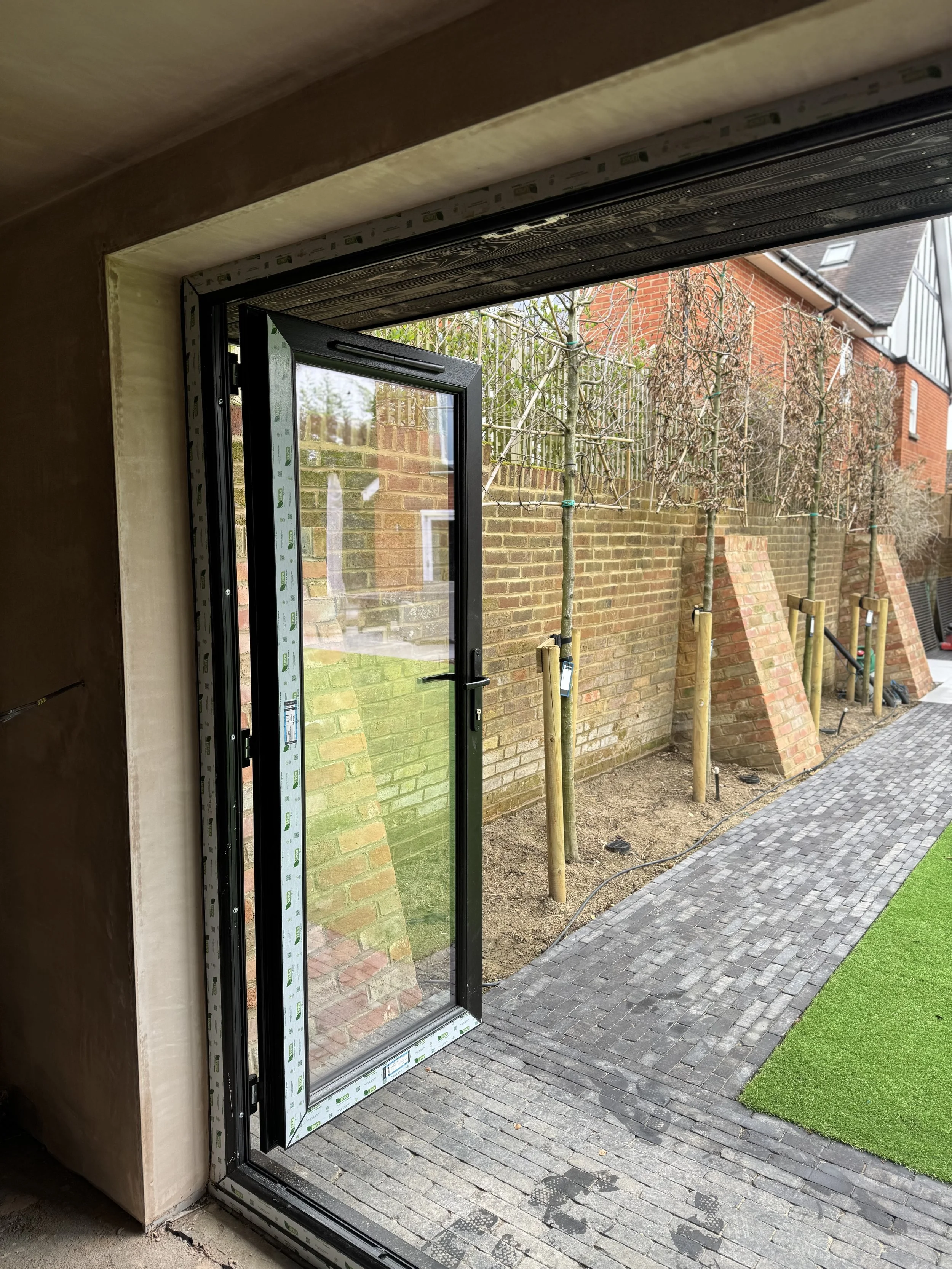 Open glass door leading to a patio with a brick wall, young trees supported by wooden stakes, a cobblestone pathway, and a lawn.