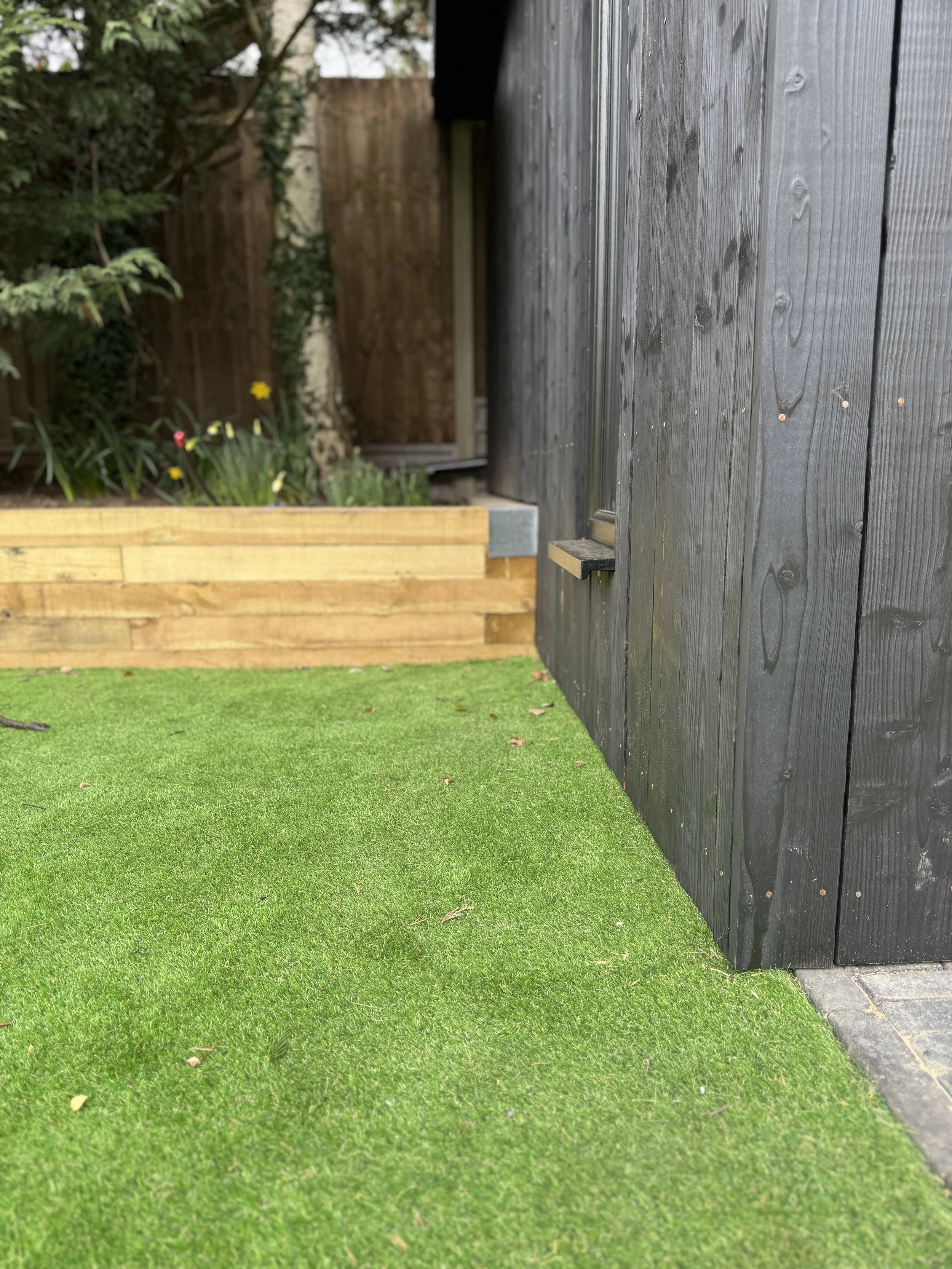 A corner of a backyard with green artificial grass, a black wooden wall, a wooden retaining wall with plants, and a bamboo fence in the background.