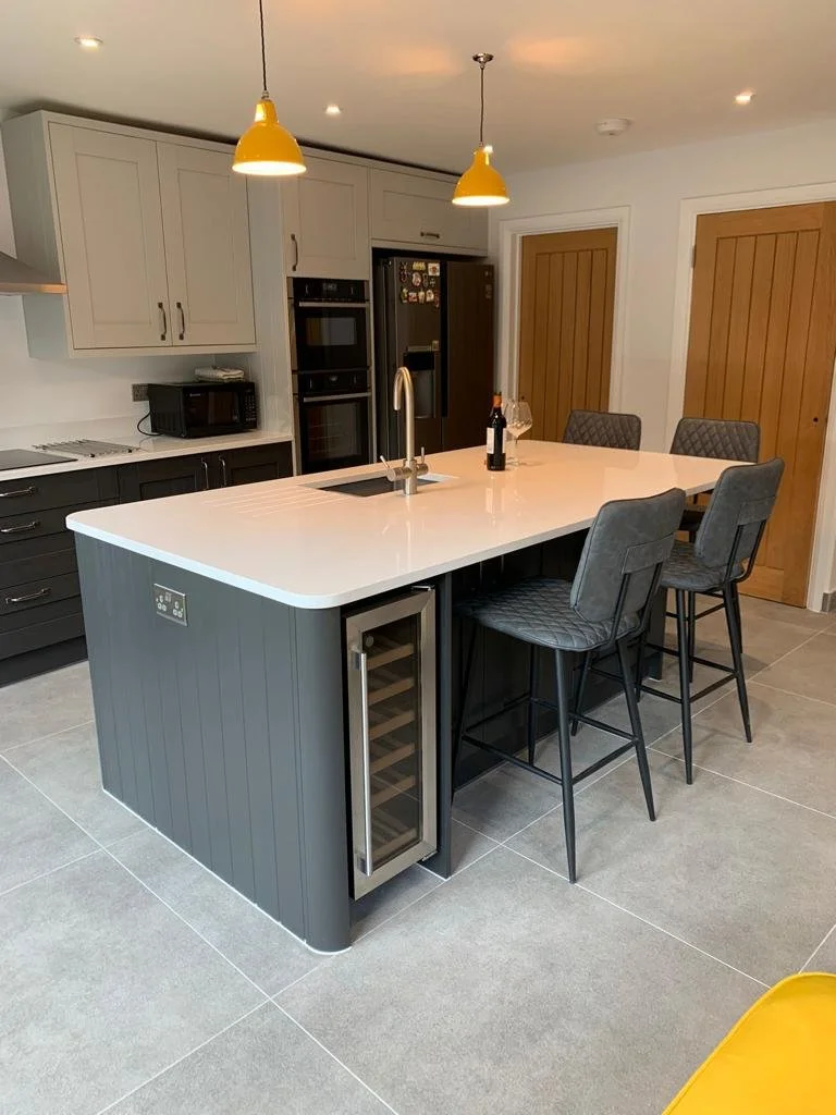 Modern kitchen with a large white island countertop, four gray bar stools, yellow pendant lights, and wood-paneled doors.