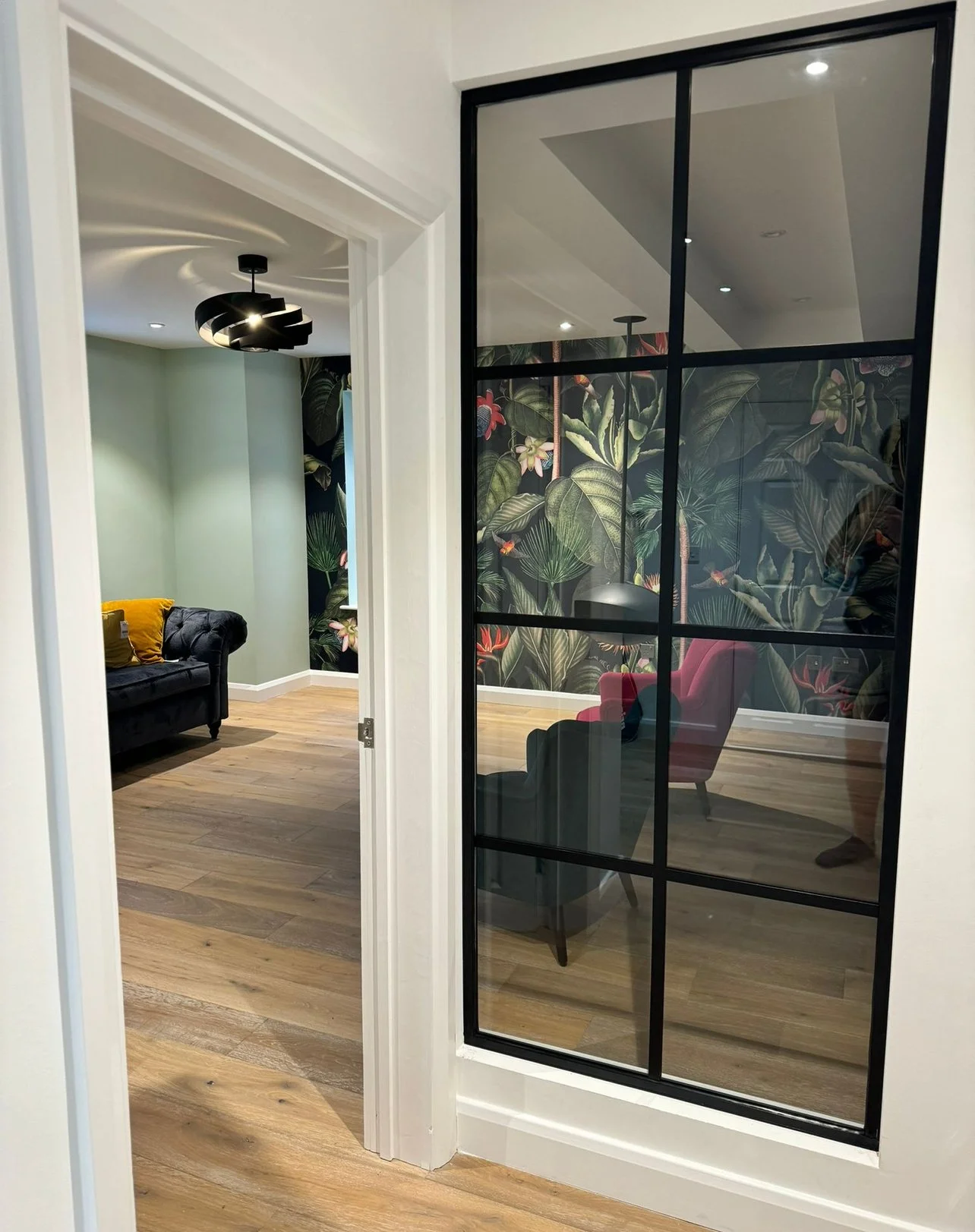 Interior view of a home with a black glass pane door, wooden flooring, and a botanical wallpapered wall. The room on the left has a dark blue tufted sofa with a yellow pillow, and a black modern ceiling light fixture. The room on the right contains a