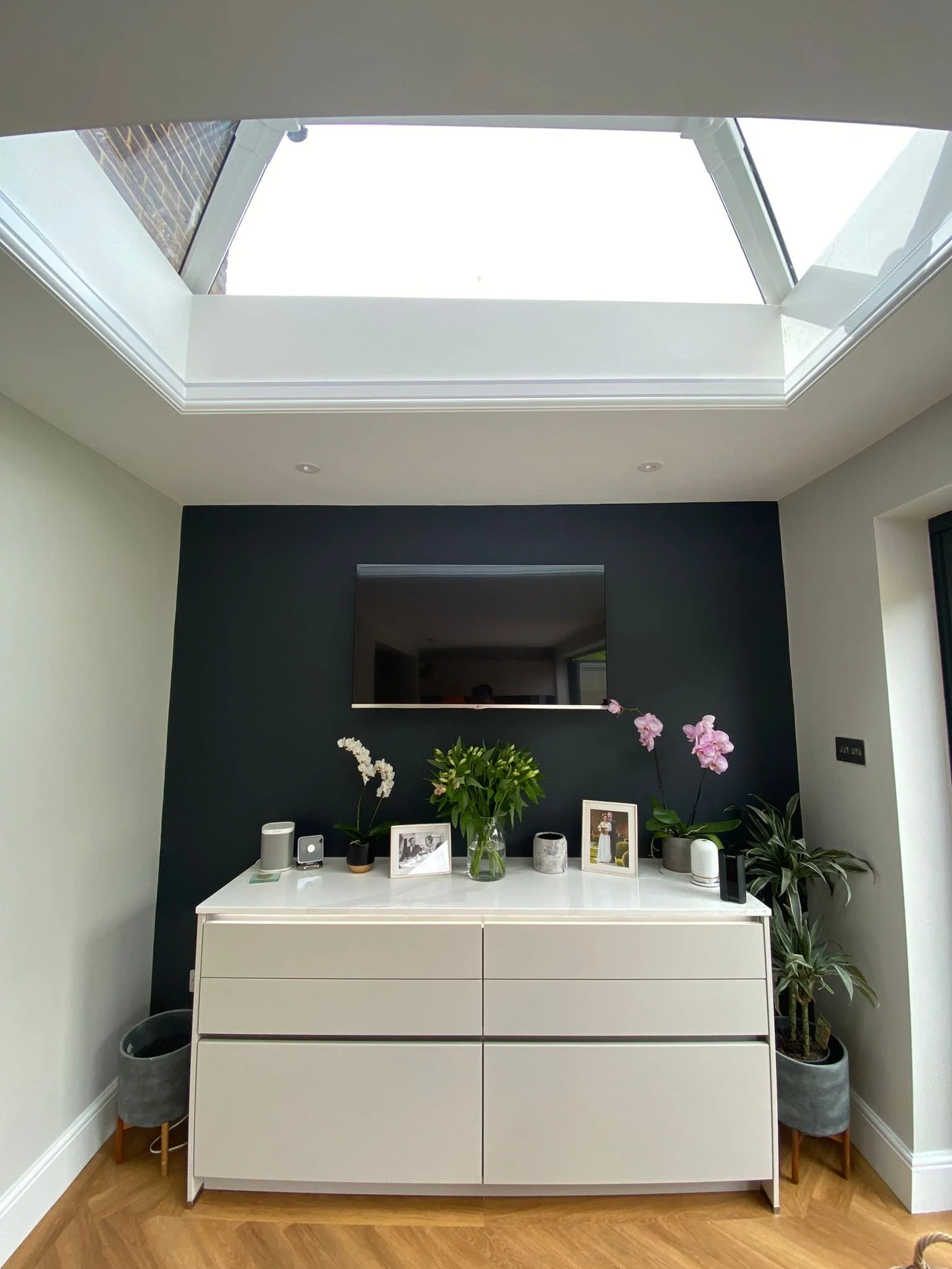 Modern interior room with a skylight, white dresser with plants and framed photos, and a dark accent wall.