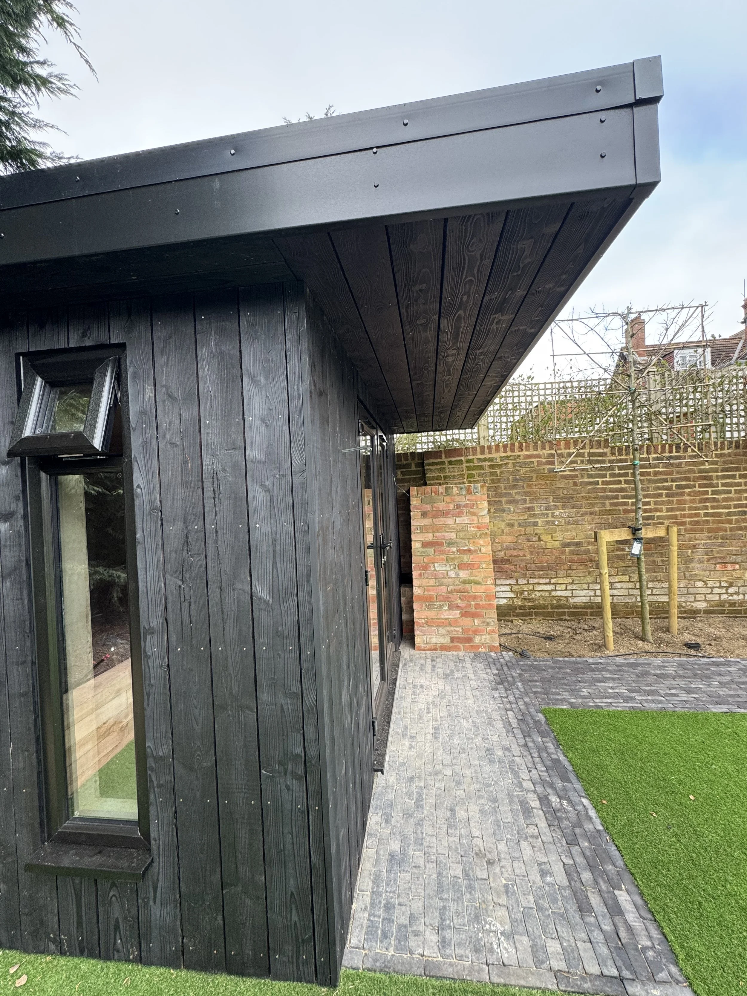 Exterior corner of a modern black wooden building with a small window, brick wall, paved walkway, artificial grass, and a young tree in a protective wooden frame.