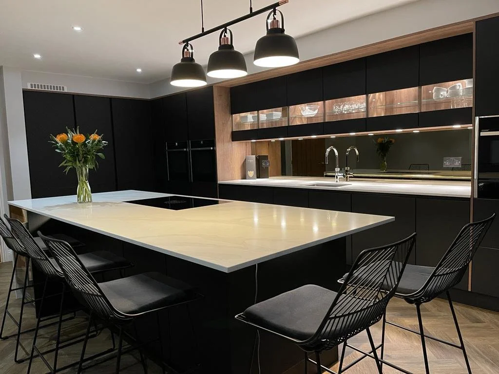 Modern kitchen with black cabinets, a large light-colored island, pendant lights, and a vase of orange flowers.