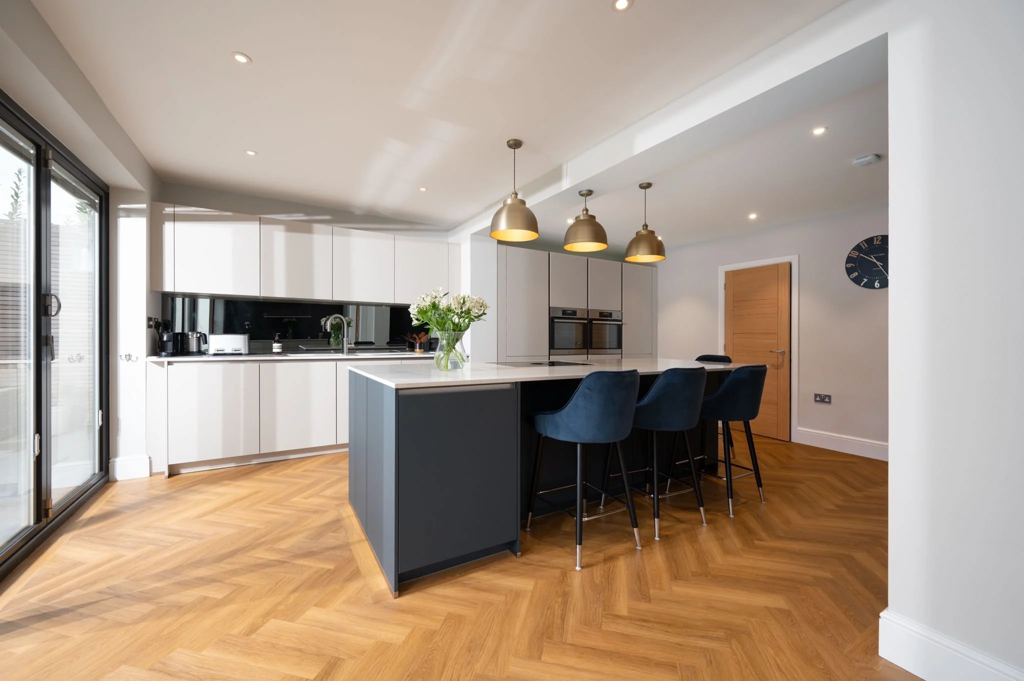 Modern kitchen with white cabinets, black backsplash, gold pendant lights, a kitchen island with a vase of white flowers, and navy blue barstools.