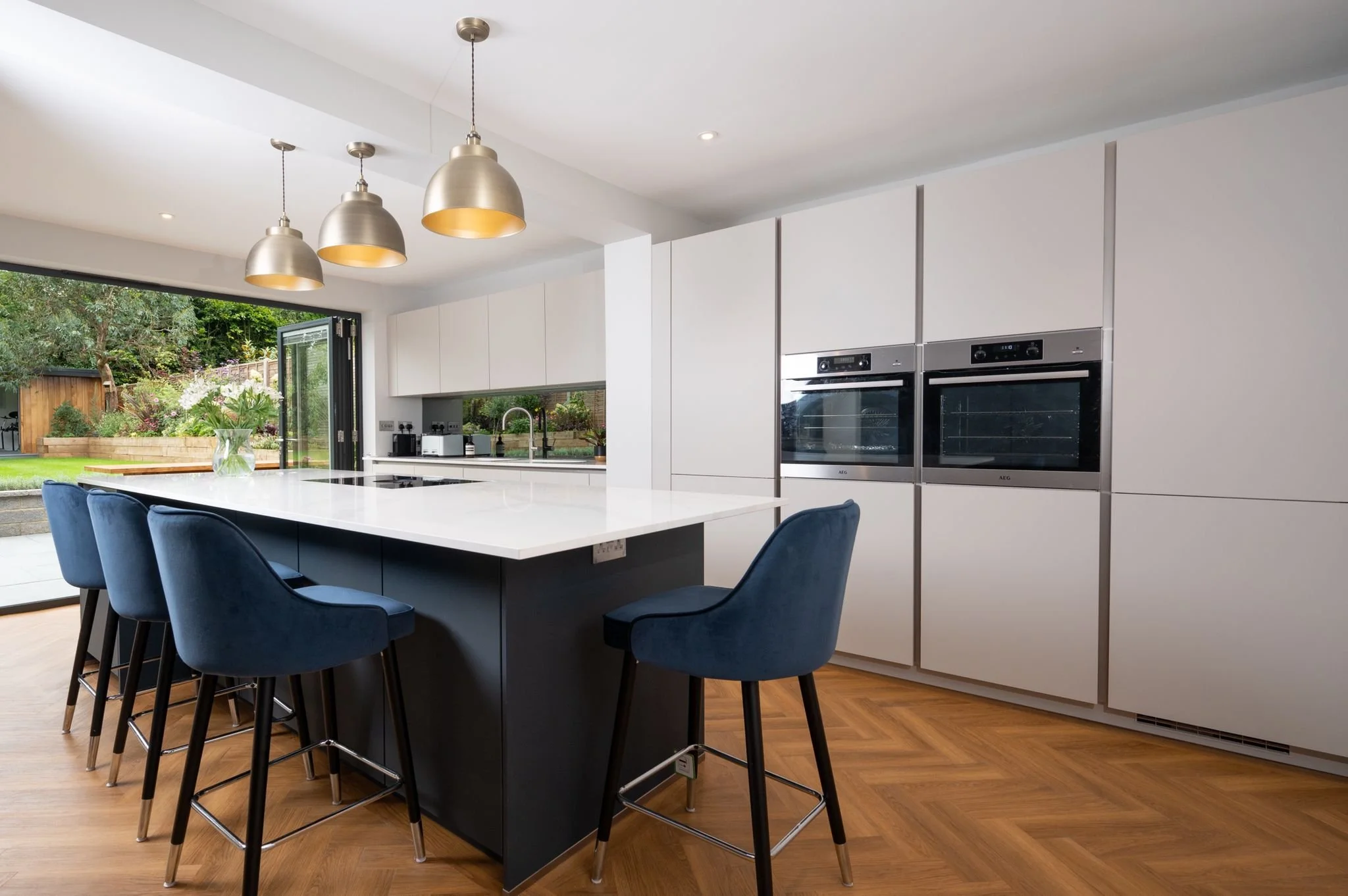 Modern kitchen with white cabinetry, a large white island with black base, blue bar stools, built-in double ovens, hardwood flooring, and large glass doors opening to a garden.