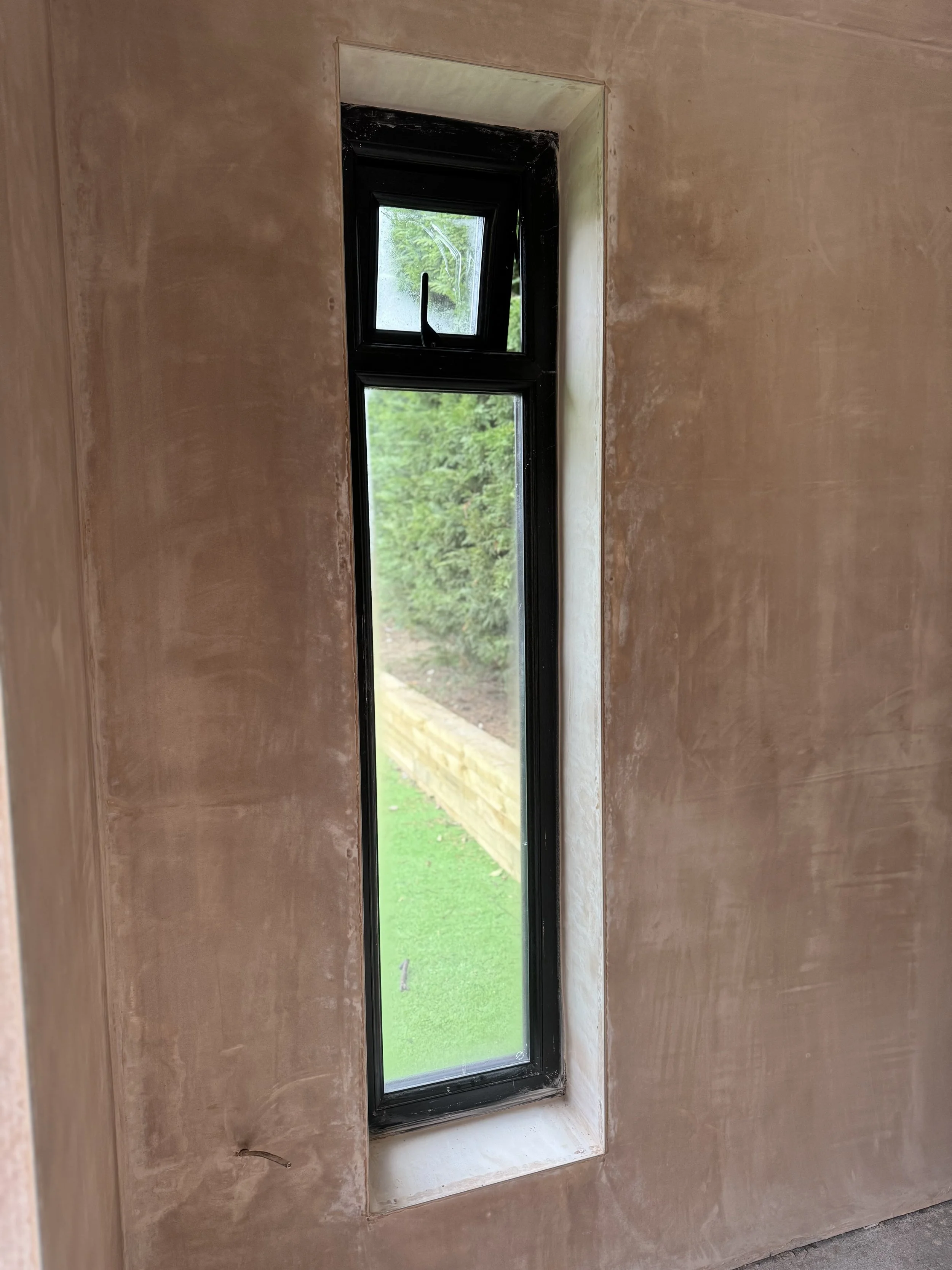 Close-up of a narrow, vertical sash window with black frame, installed in a wall with unfinished beige plaster, showing a view of green grass, a wooden border, and bushes outside.