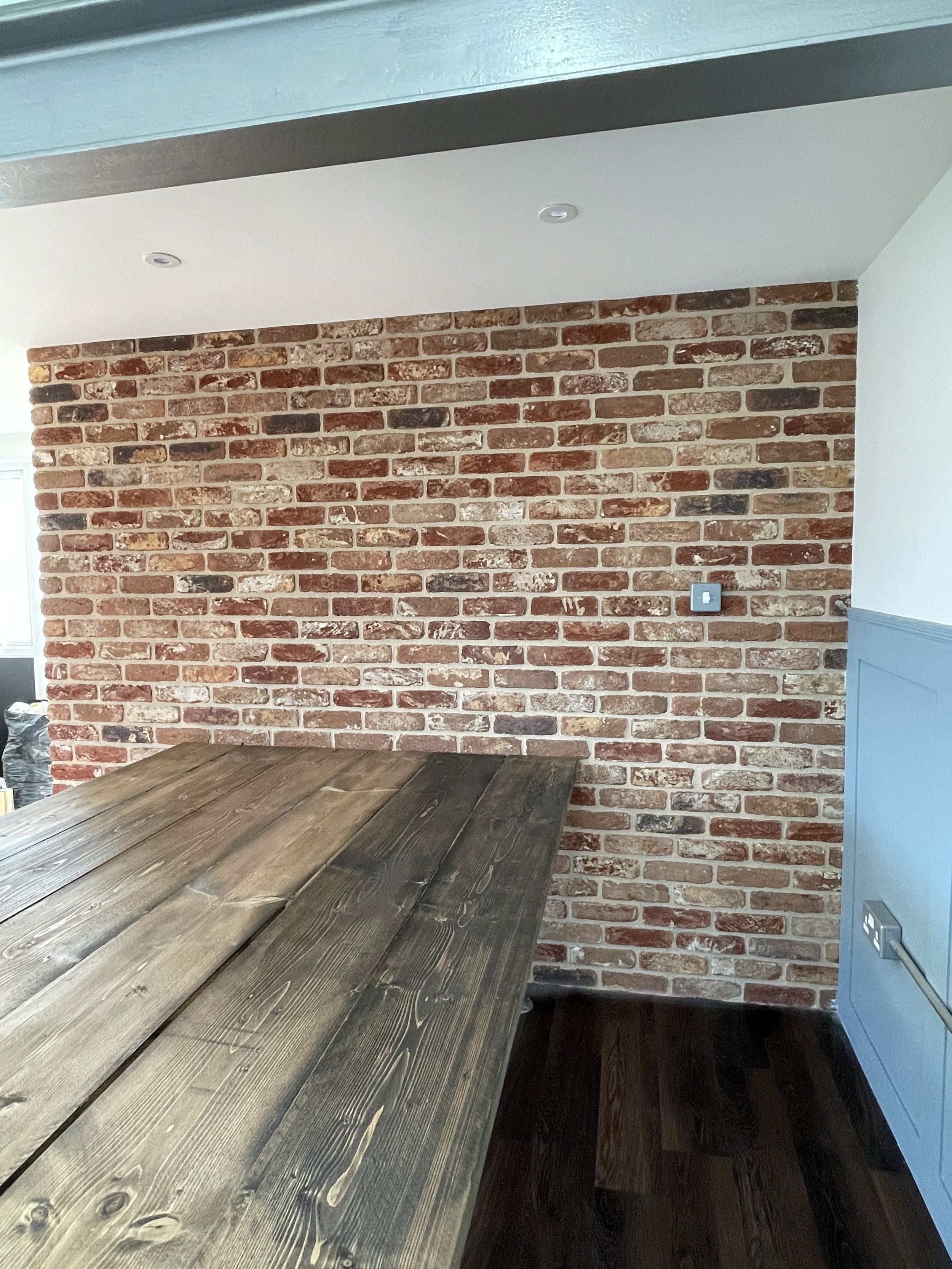 Interior space with a rustic brick wall, wooden table, and a corner with blue paneling and electrical outlets.