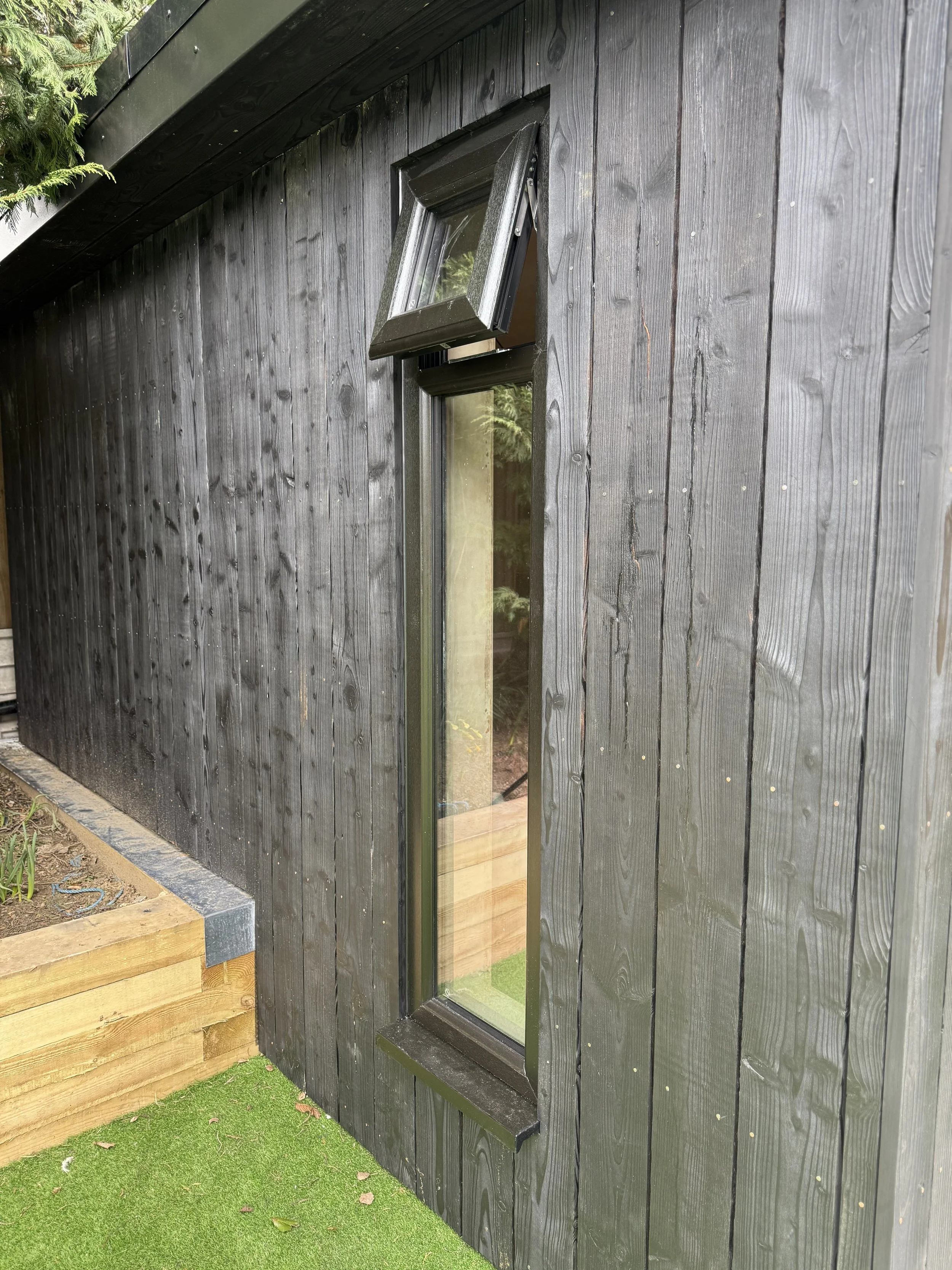 A vertical black-framed window with a small, open, awning-style window above it on the exterior of a black wooden building. The building has a black wooden wall with vertical planks, next to a wooden planter box with soil, and a green artificial gras