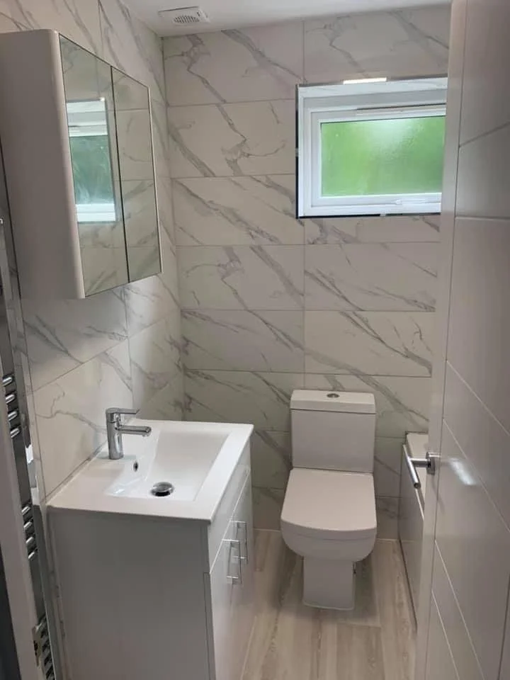 Small modern bathroom with marble walls, a white toilet, a small white sink with a cabinet, a mirror cabinet, a window with green scenery outside, and a towel rack.