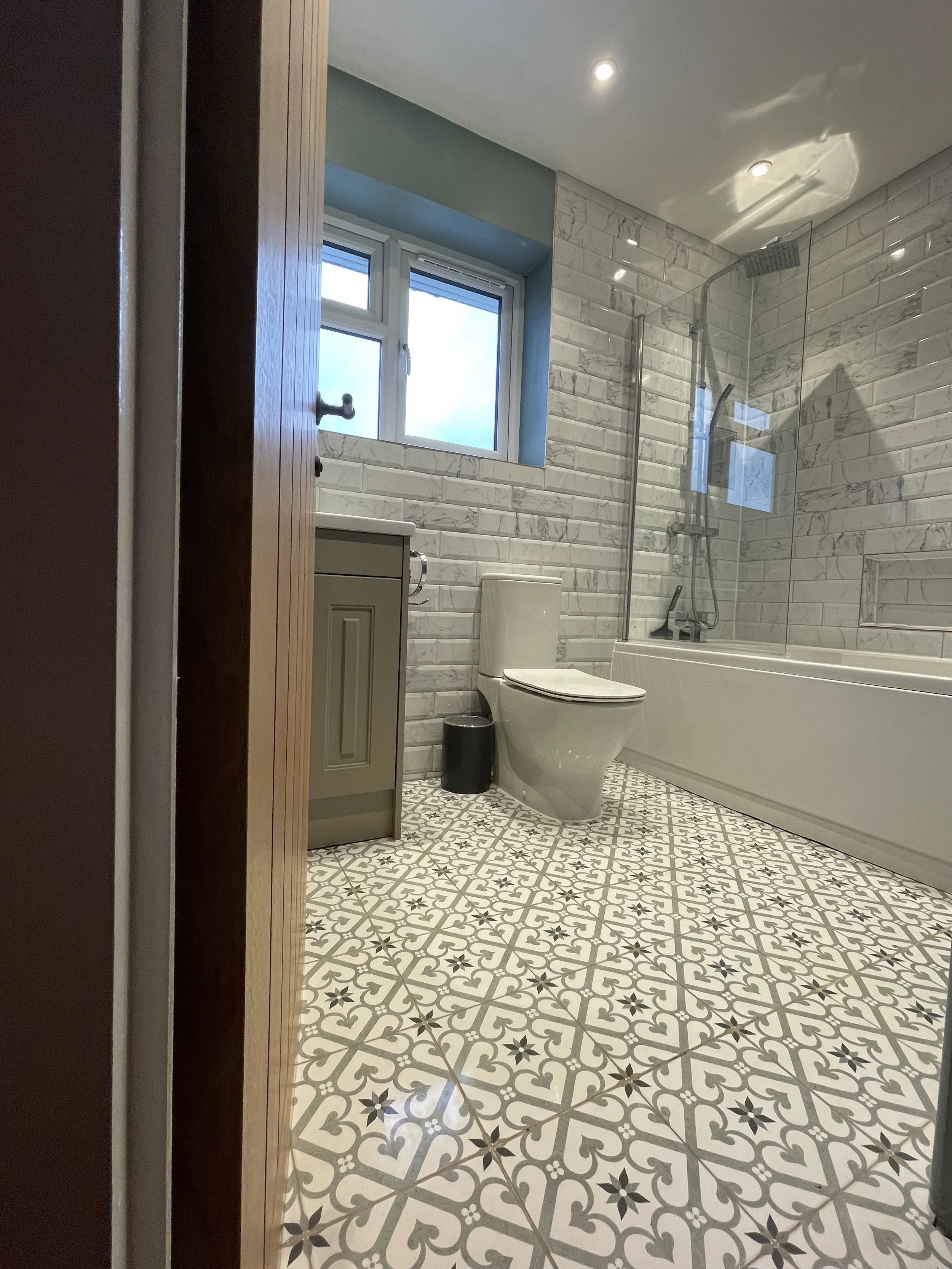 Modern bathroom with patterned floor tiles, white toilet, green vanity, bathtub with shower, window, and marble-veneer walls.