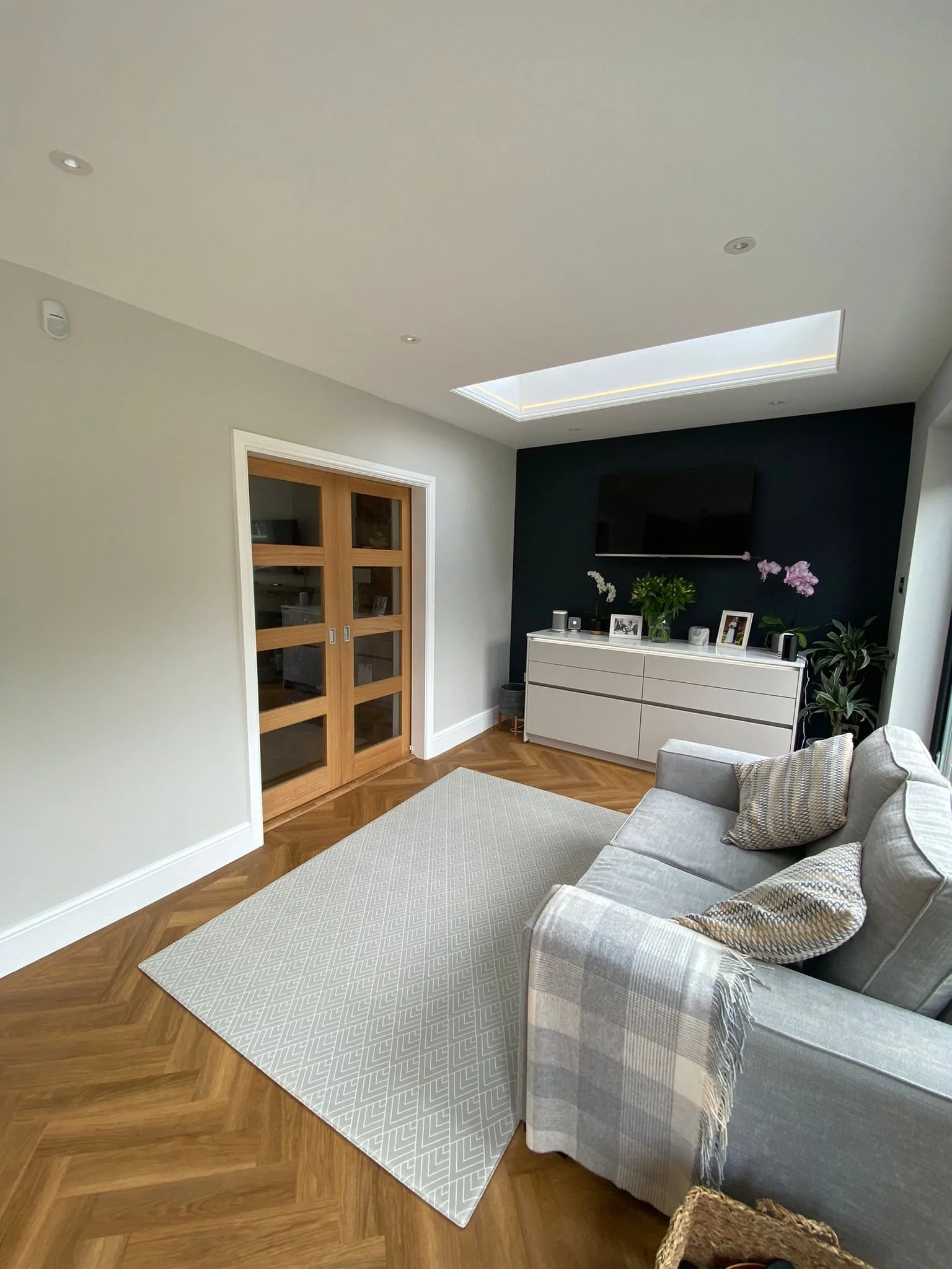 Modern living room with a gray sofa, patterned pillows, a beige rug, a half-moon white sideboard decorated with picture frames and potted plants, a black accent wall with a mounted TV, and a skylight in the ceiling.