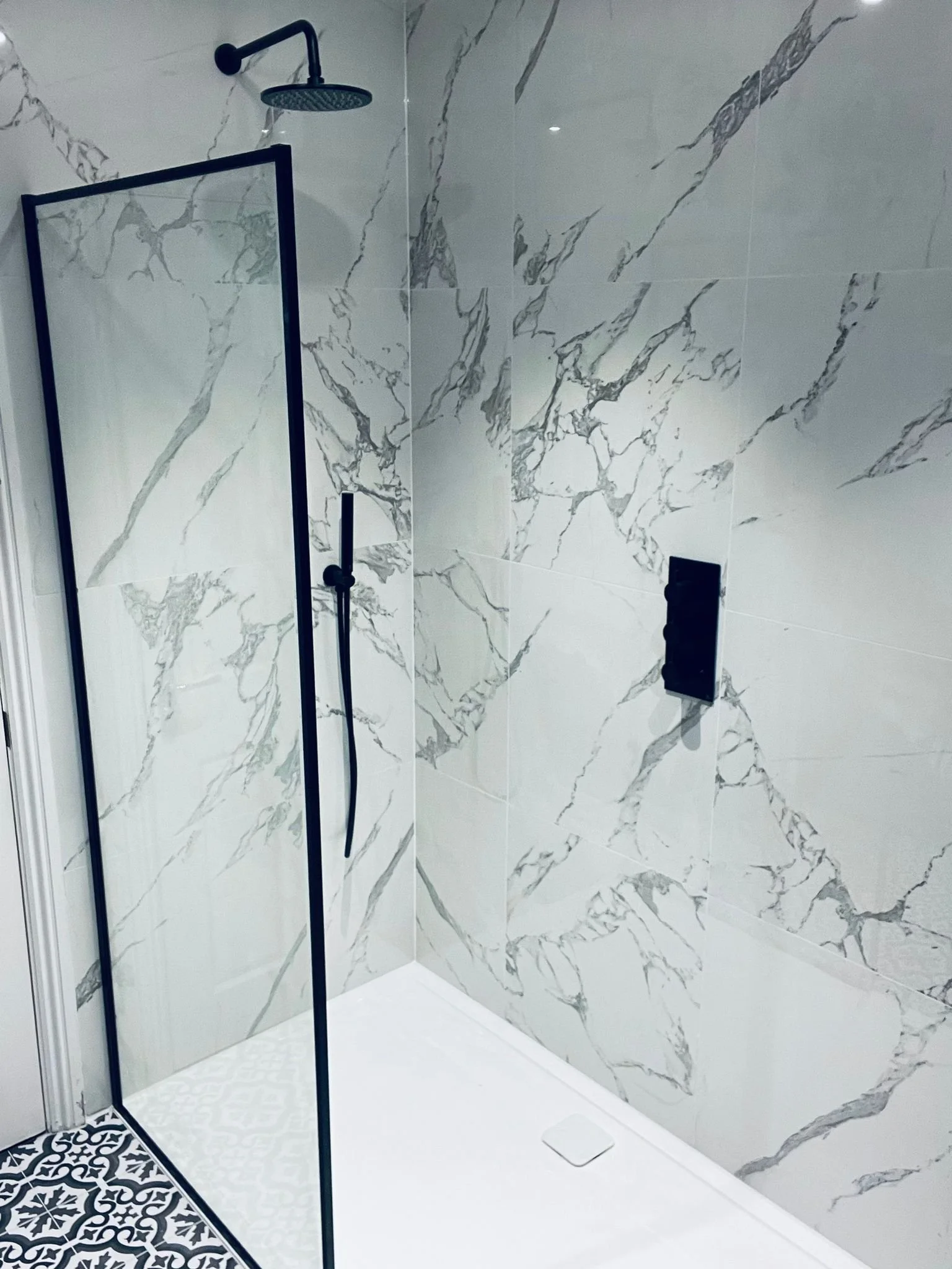 Modern walk-in shower with large marble tiles, a black showerhead, a black handheld shower, a glass divider, and decorative black and white patterned floor tiles.