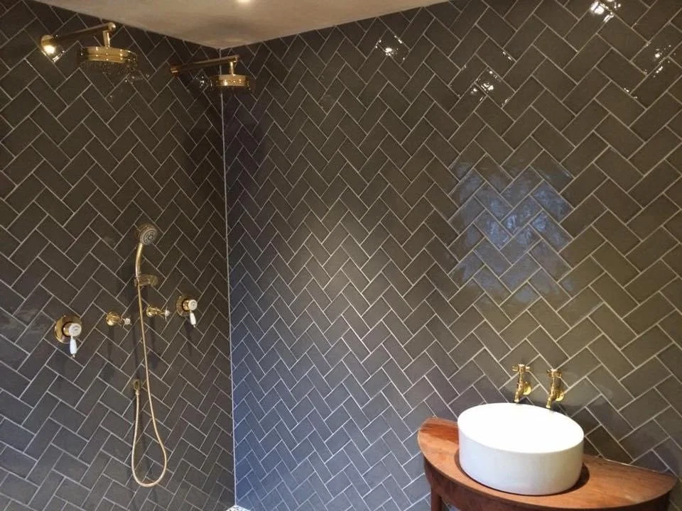 Bathroom with dark herringbone tile wall, gold shower fixtures, and a white oval vessel sink on a wooden countertop.
