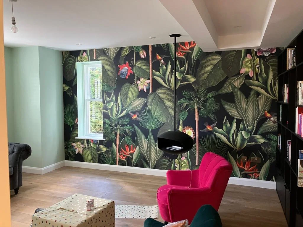 Living room with tropical leaf and bird wallpaper, red armchair, black bookshelf, and window with greenery outside.
