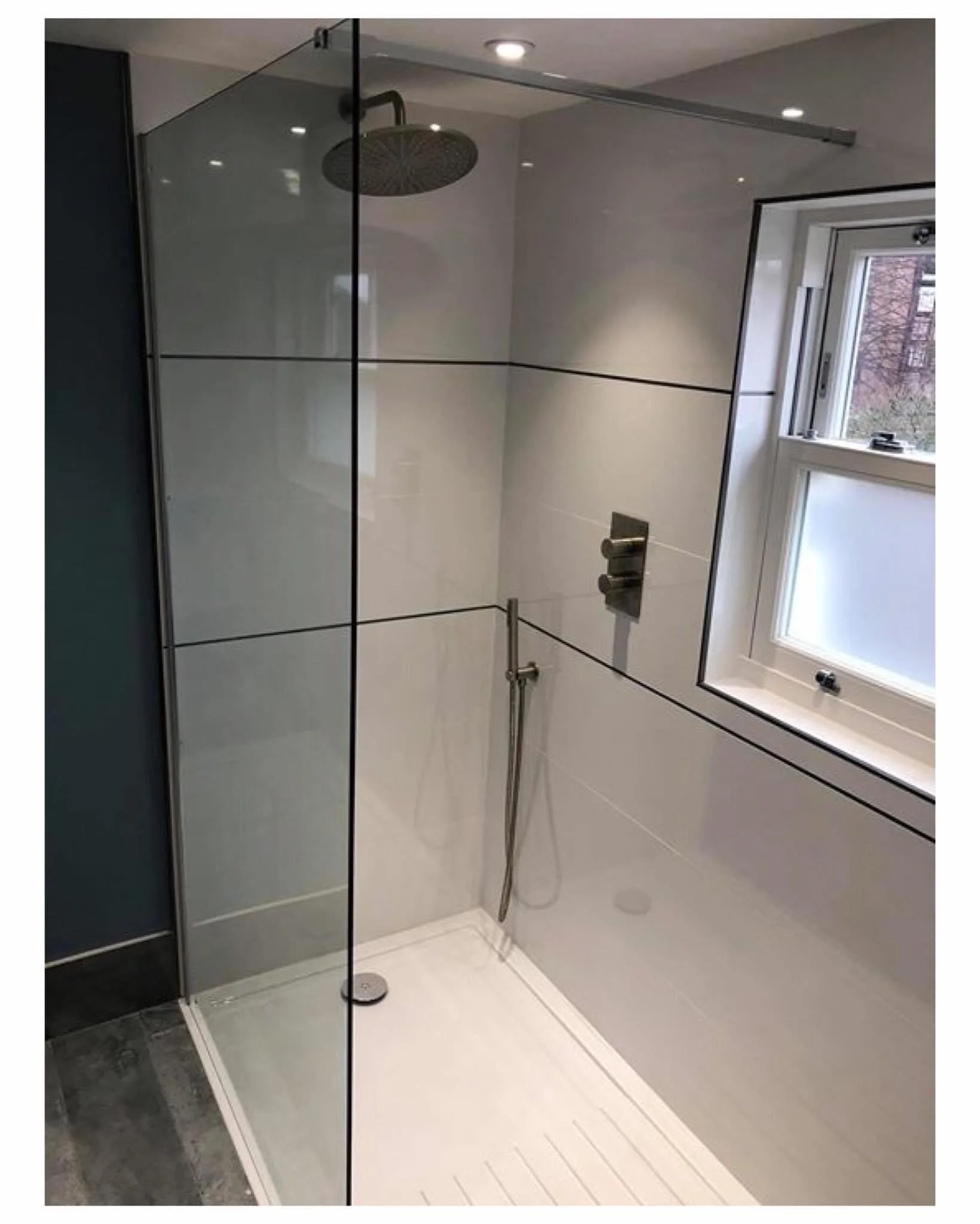 Modern shower with glass enclosure, large showerhead, hand-held shower, white tile walls, small window, and dark floor tiles.