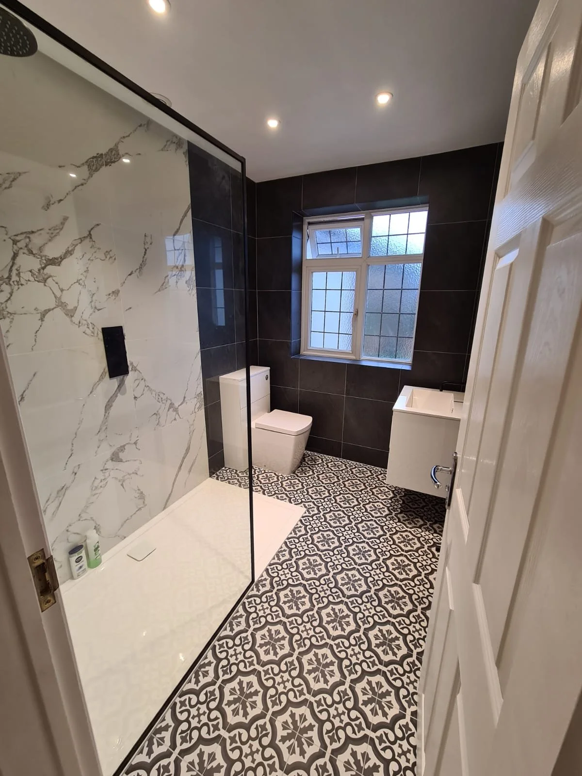 Modern bathroom with patterned floor tiles, a toilet, a small sunk-in sink, a window with frosted glass, and a walk-in shower with large marble-patterned tiles and a clear glass door.