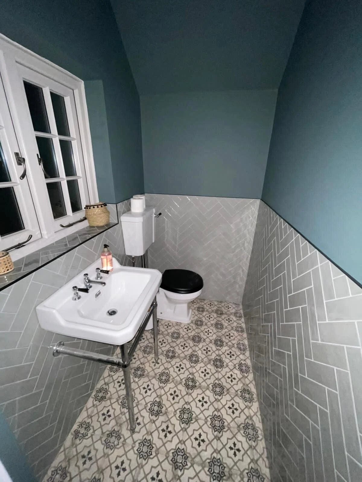 A small bathroom with a white sink, black toilet, blue walls, patterned tile floor, and a window with white trim.