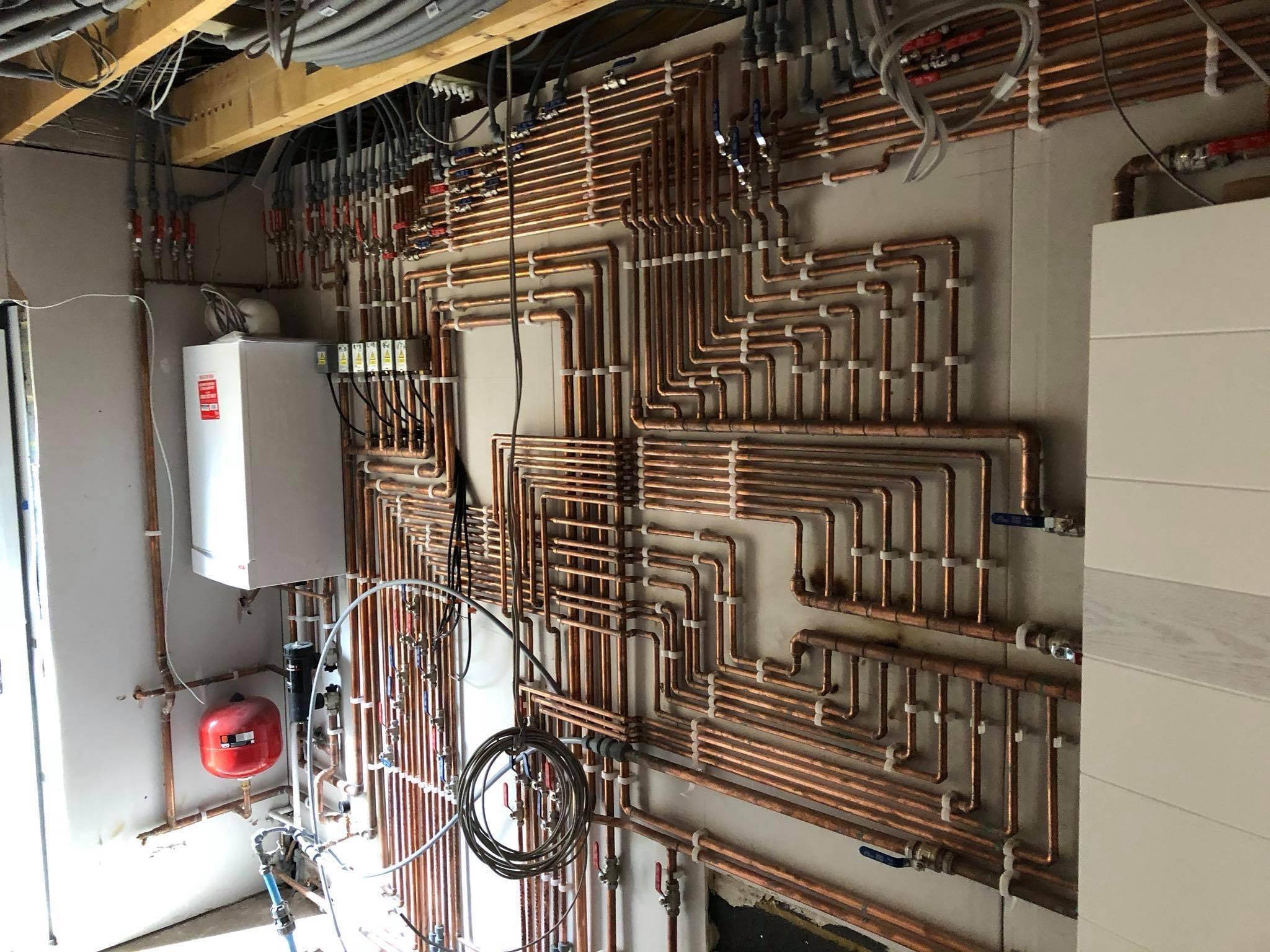 A complex network of copper plumbing pipes and electrical wiring in a utility room, with a white water heater and a red expansion tank mounted on the wall.